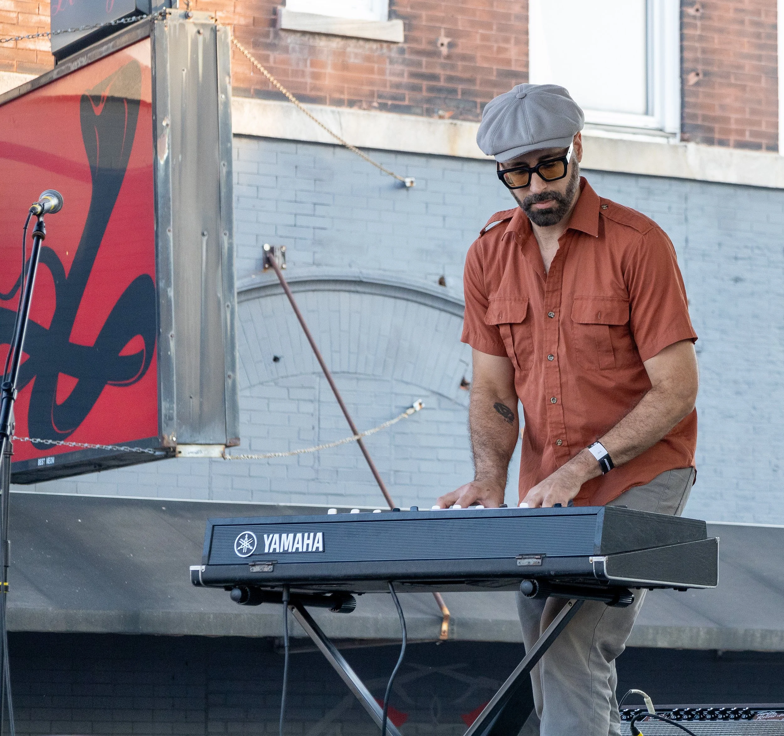 A man wearing glasses, a gray beret, and a burnt orange shirt playing a Yamaha keyboard outdoors, with brick and concrete buildings in the background.