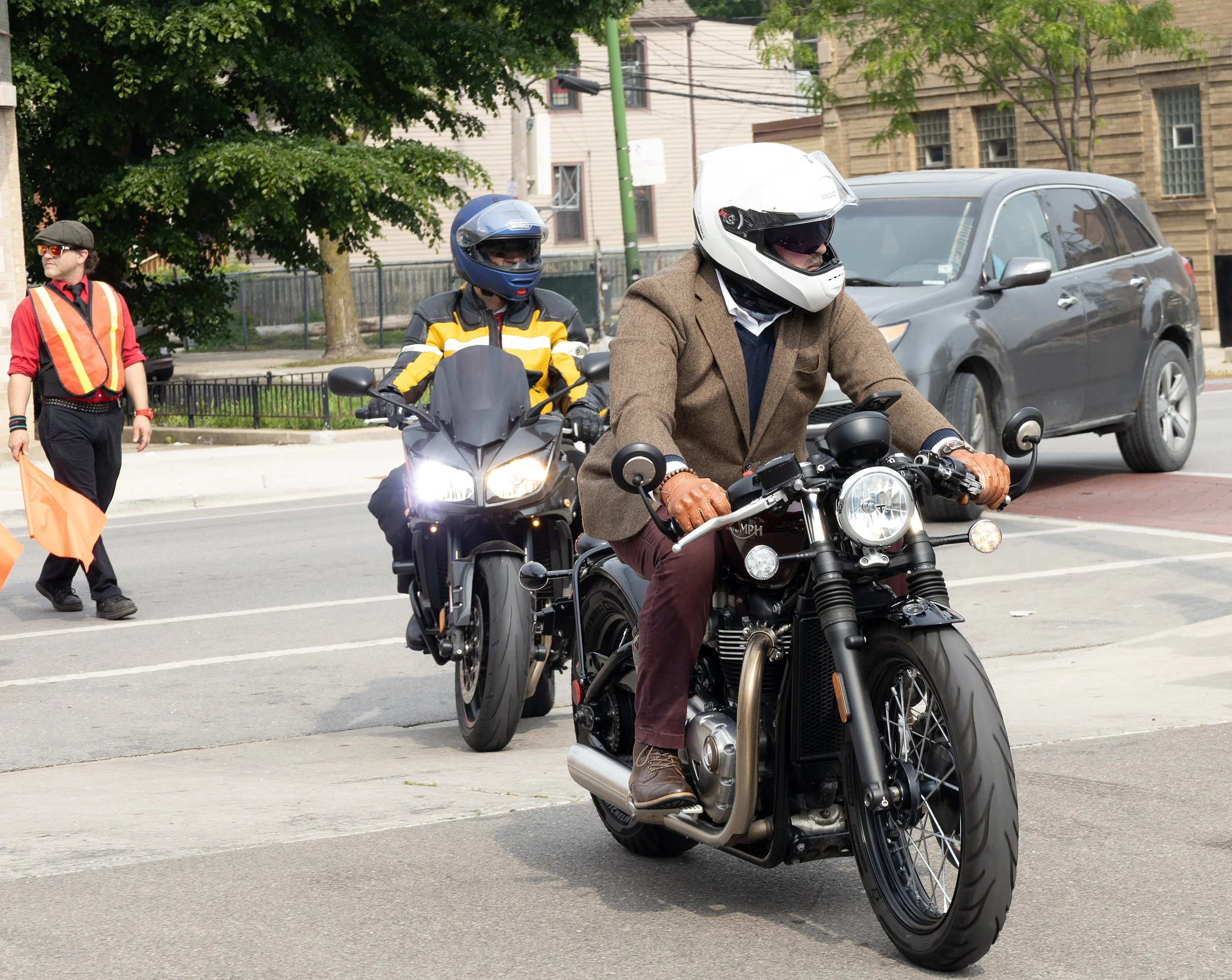 Multiple people riding and walking near parked motorcycles on a city street, with trees and buildings in the background.