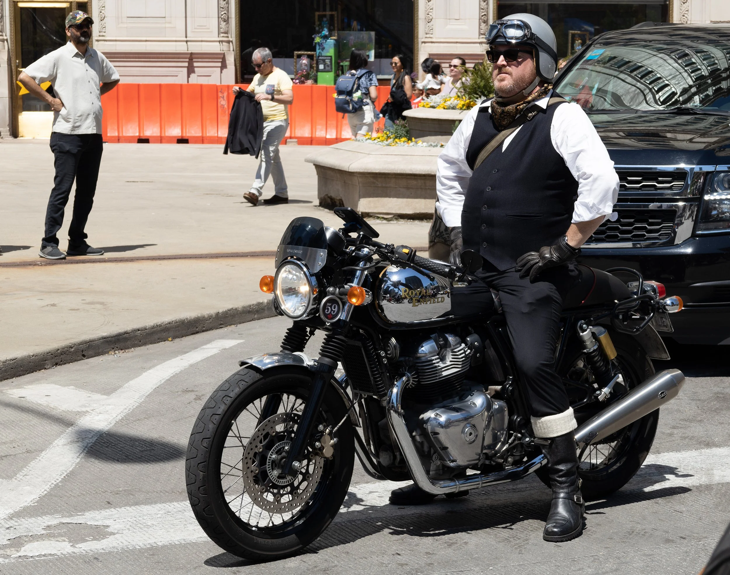 A man wearing a black vest, white shirt, black helmet with goggles, and black gloves sitting on a black Royal Enfield motorcycle on the street, with a city scene and several pedestrians in the background.