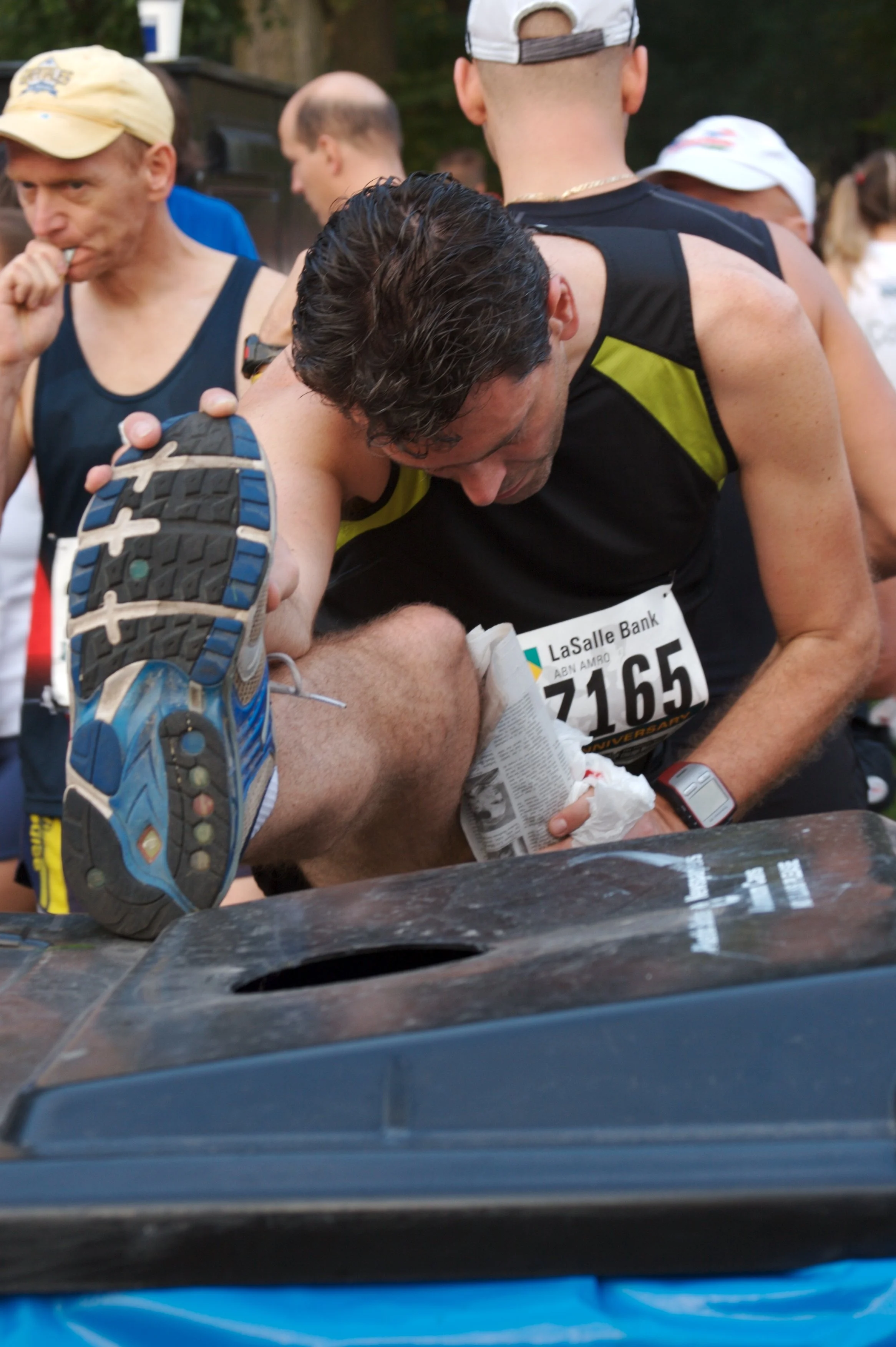 A group of runners at the finish line of a race, with one man helping another person who appears to be exhausted. The runners are wearing athletic clothing and race bibs, and there is a crowd in the background.