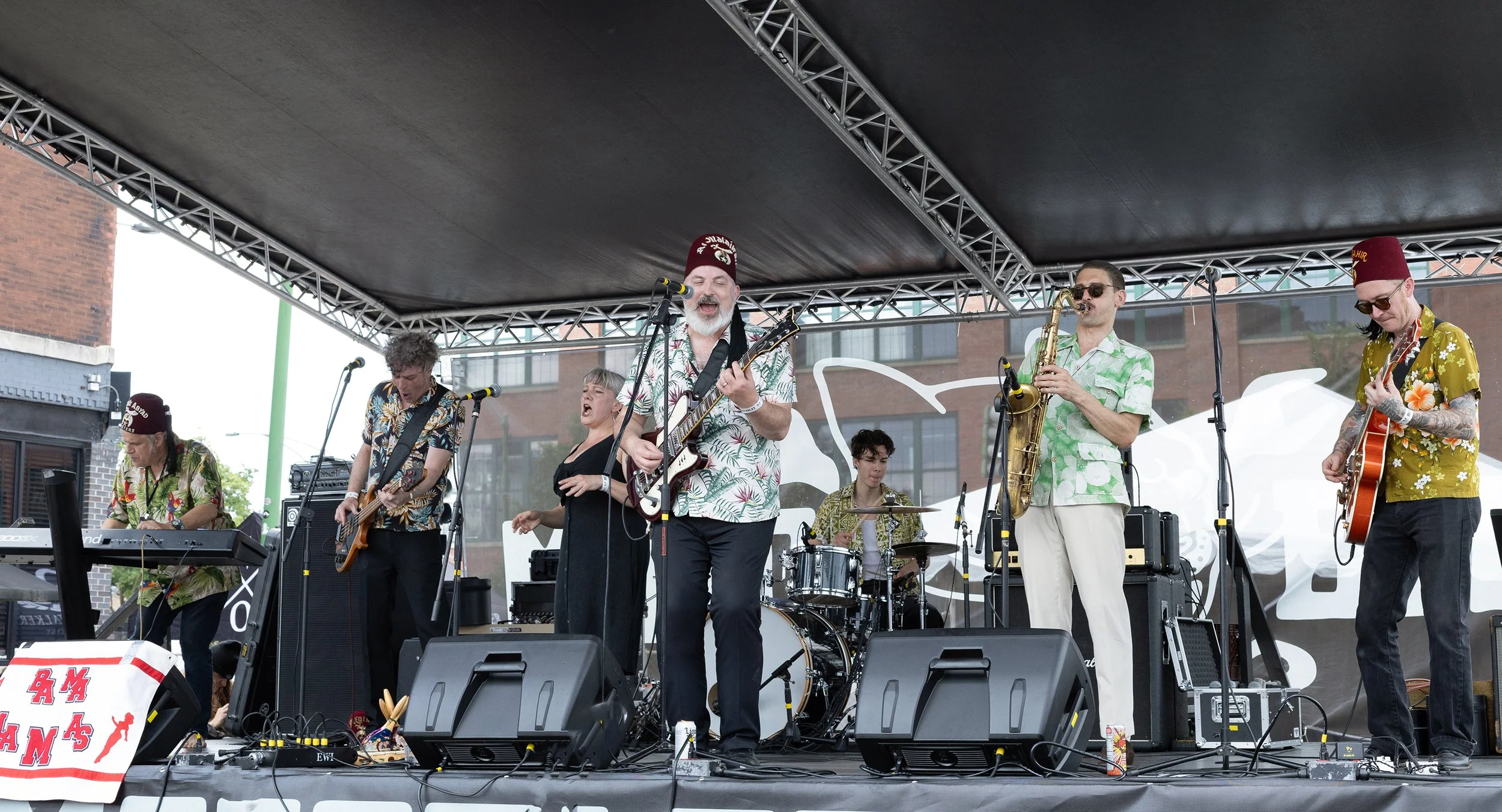 A band performing on an outdoor stage with six members playing various instruments and singing, dressed in colorful tropical shirts and hats, under a black canopy.