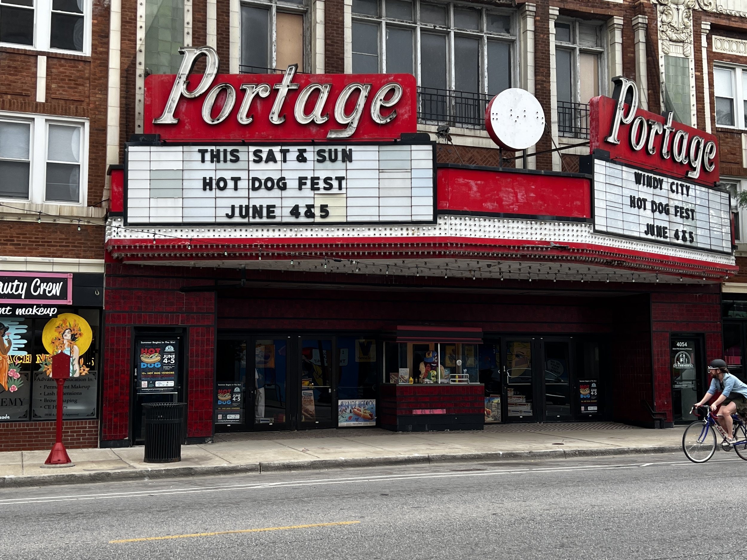 The image shows the exterior of a theater or venue called the Portage with a marquee advertising a Hot Dog Fest event happening on June 4th and 5th on Saturday and Sunday. There is a person riding a bicycle on the street in front of the venue, and a 