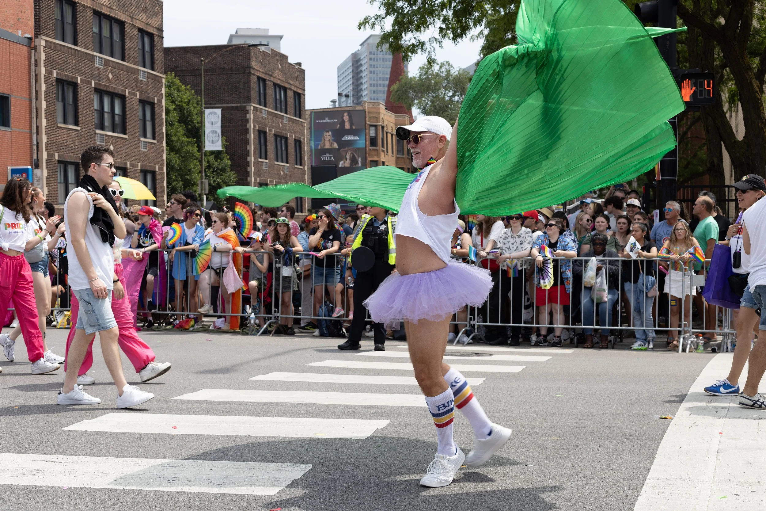 A person with a white baseball cap, sunglasses, rainbow-striped socks, and a purple tutu, holding a large green parasol, walking in a parade or festival on a city street with a crowd of onlookers behind a barrier.