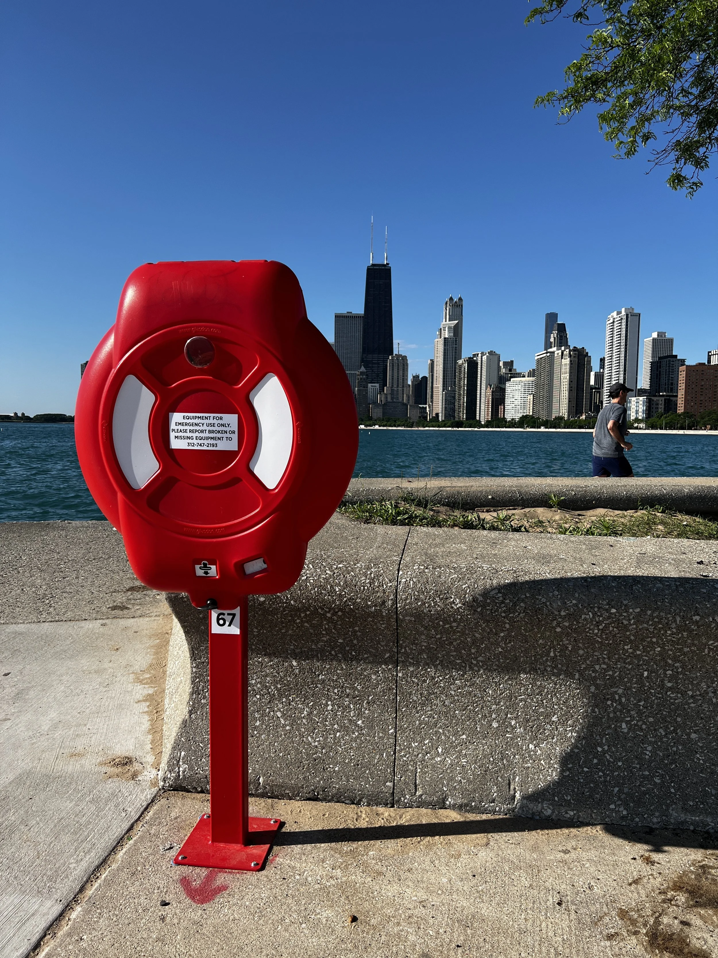 Red emergency life ring station on sidewalk with city skyline in the background, including tall skyscrapers and a person walking along the water.