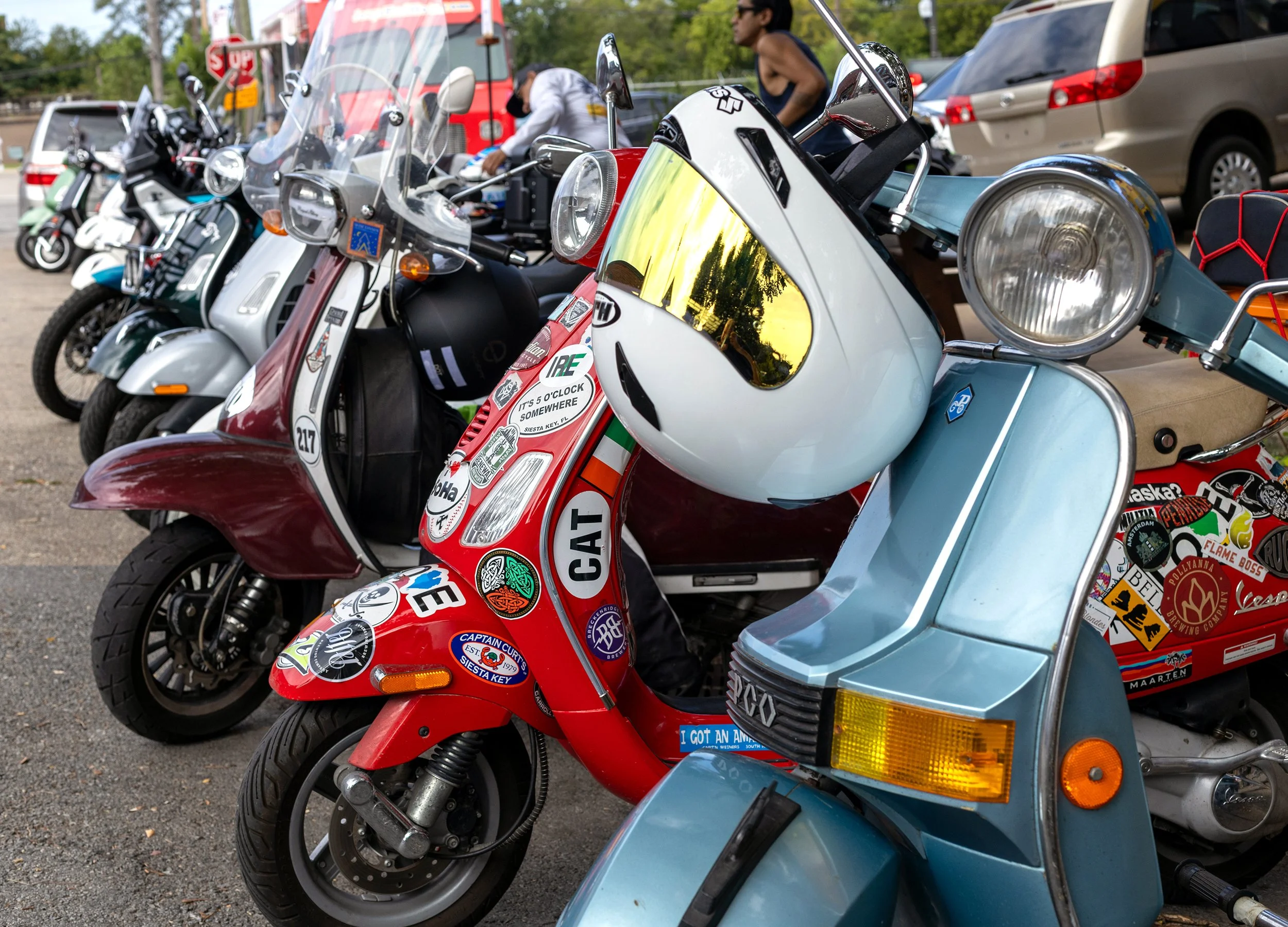 Multiple scooters parked in a parking lot, with a person in the background working on something and another person standing nearby. There are cars visible in the background and stickers on the scooters.