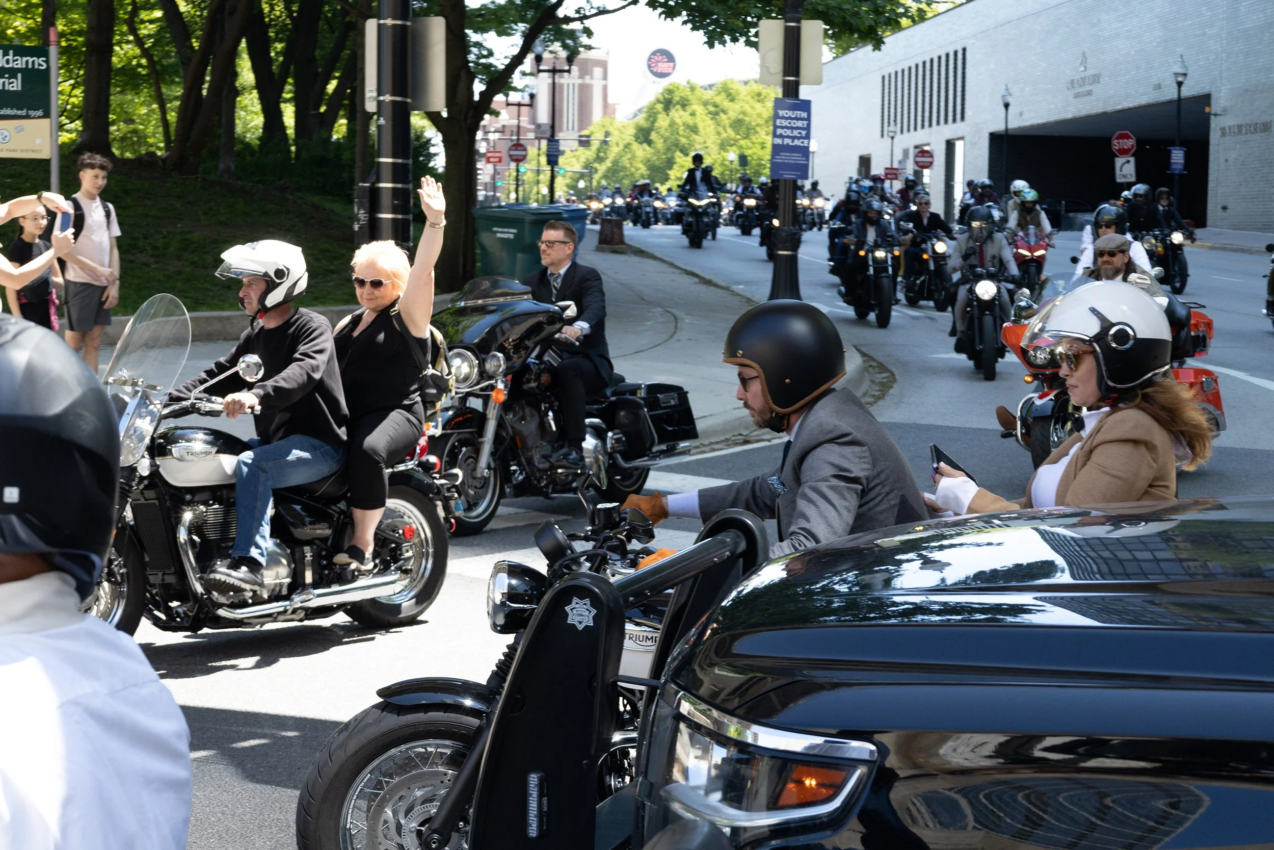 A parade of motorcyclists riding on a city street, with some waving at the crowd. The scene includes spectators, street signs, and a modern building in the background.
