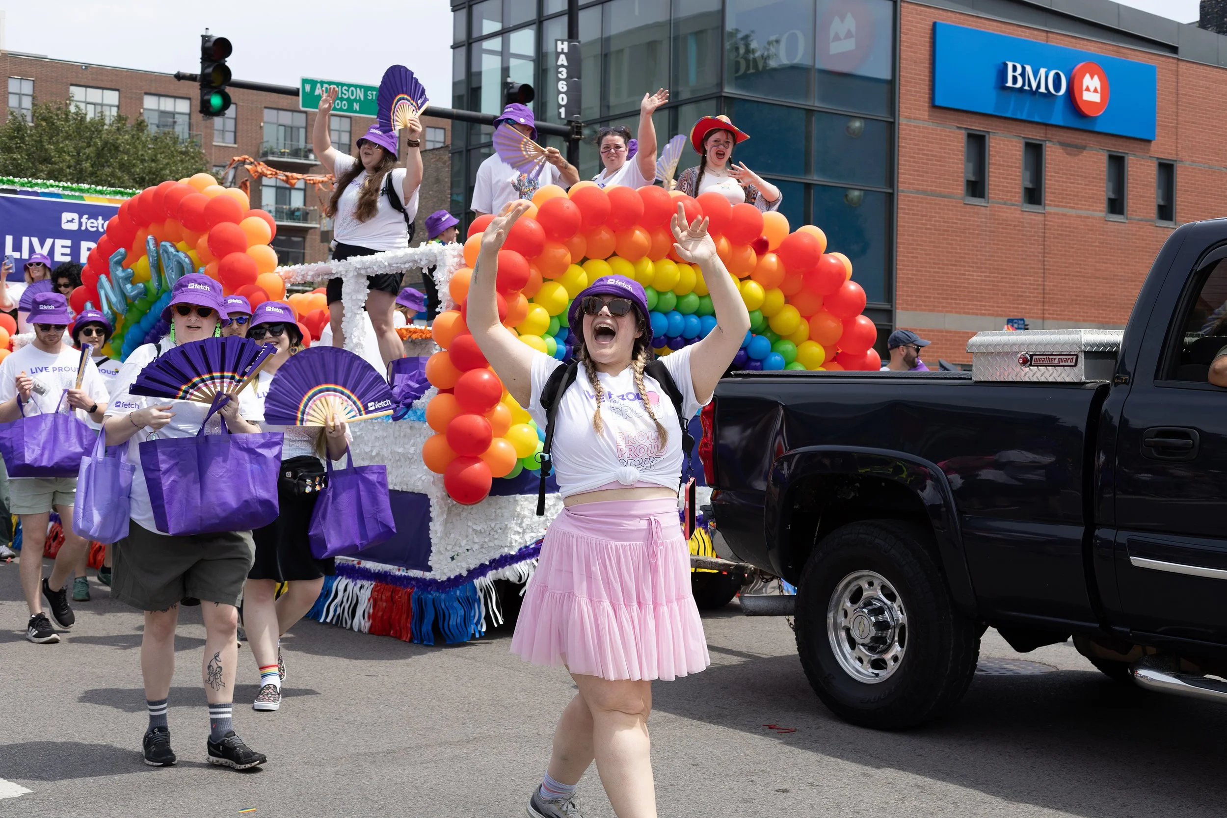 A lively parade with people walking and dancing, including a woman in a pink skirt and braids, surrounded by a float decorated with rainbow balloons and people wearing purple hats and shirts, with a BMO bank building in the background.