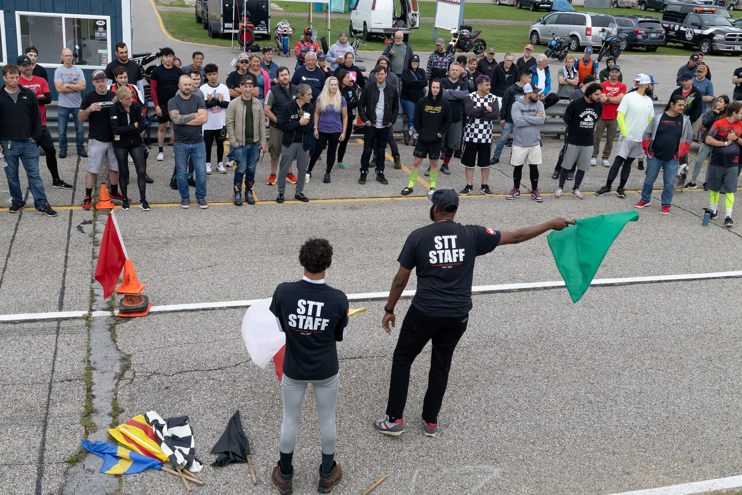 A large group of people gathered outdoors at a motorcycle event, standing behind a line marked by traffic cones. A man in a black shirt with 'STT STAFF' on the back is coordinating. The crowd includes diverse individuals, some wearing casual attire a
