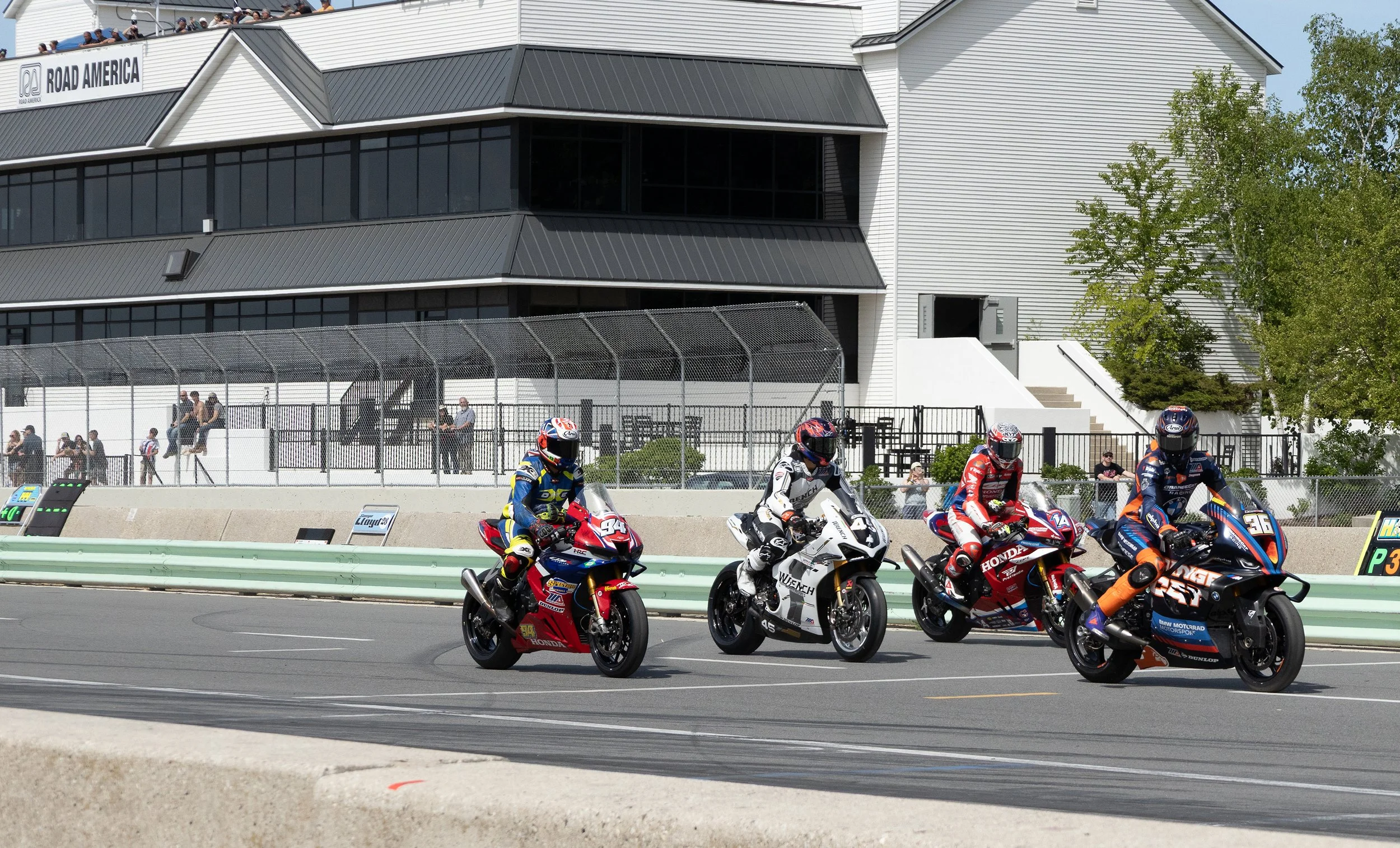 Motorcycle racers in race gear riding on a racetrack, with spectators watching from the stands behind a fence and a large building in the background.