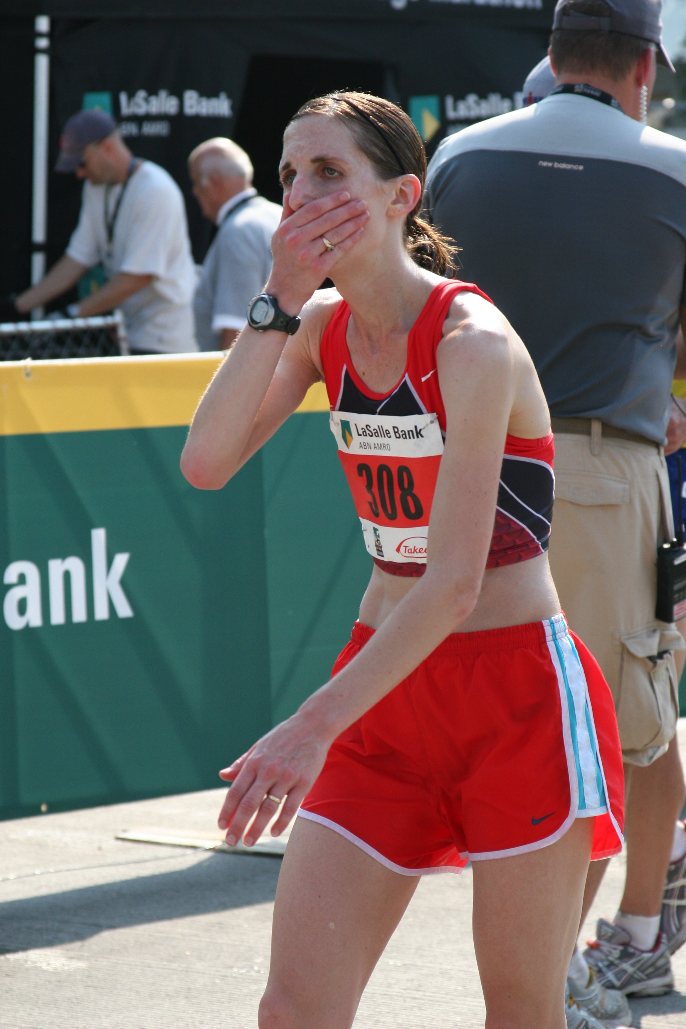 A female athlete wearing a red running uniform with the number 308, standing at a race event, covering her mouth with her hand, with other people and a LaSalle Bank banner in the background.