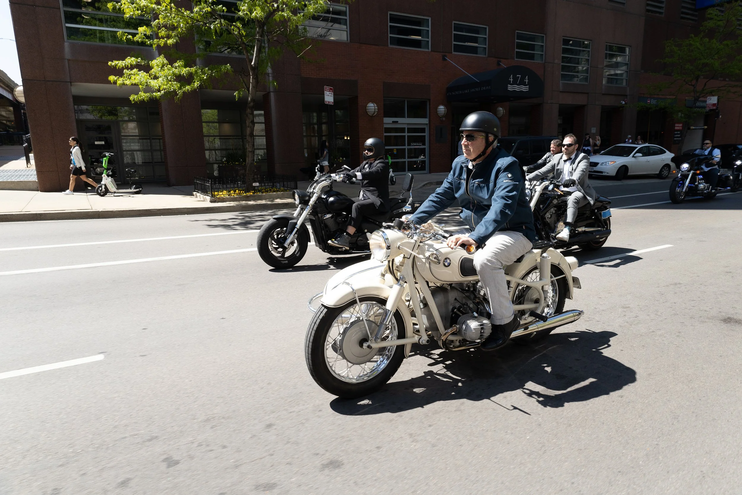 Multiple motorcyclists riding on a city street near a brick building with glass windows, with pedestrians and parked cars visible in the background, and a woman walking alongside riding an electric scooter.