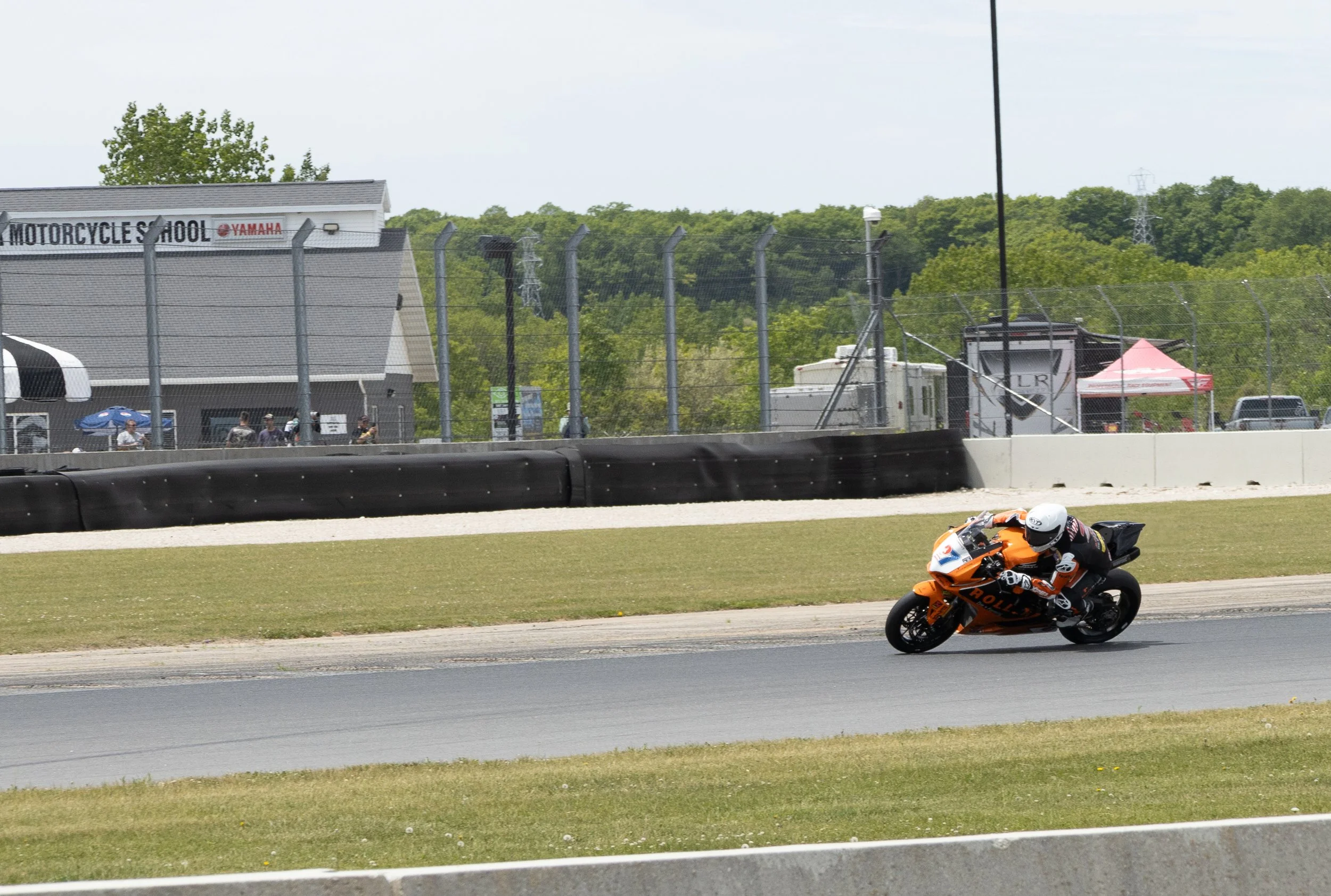 Motorcycle racing on a track, with a rider wearing a white helmet and orange racing suit, leaning into a turn on an orange Kawasaki motorcycle. In the background, there is a fence, a building with a sign that reads 'Motorcycle School' with Yamaha log