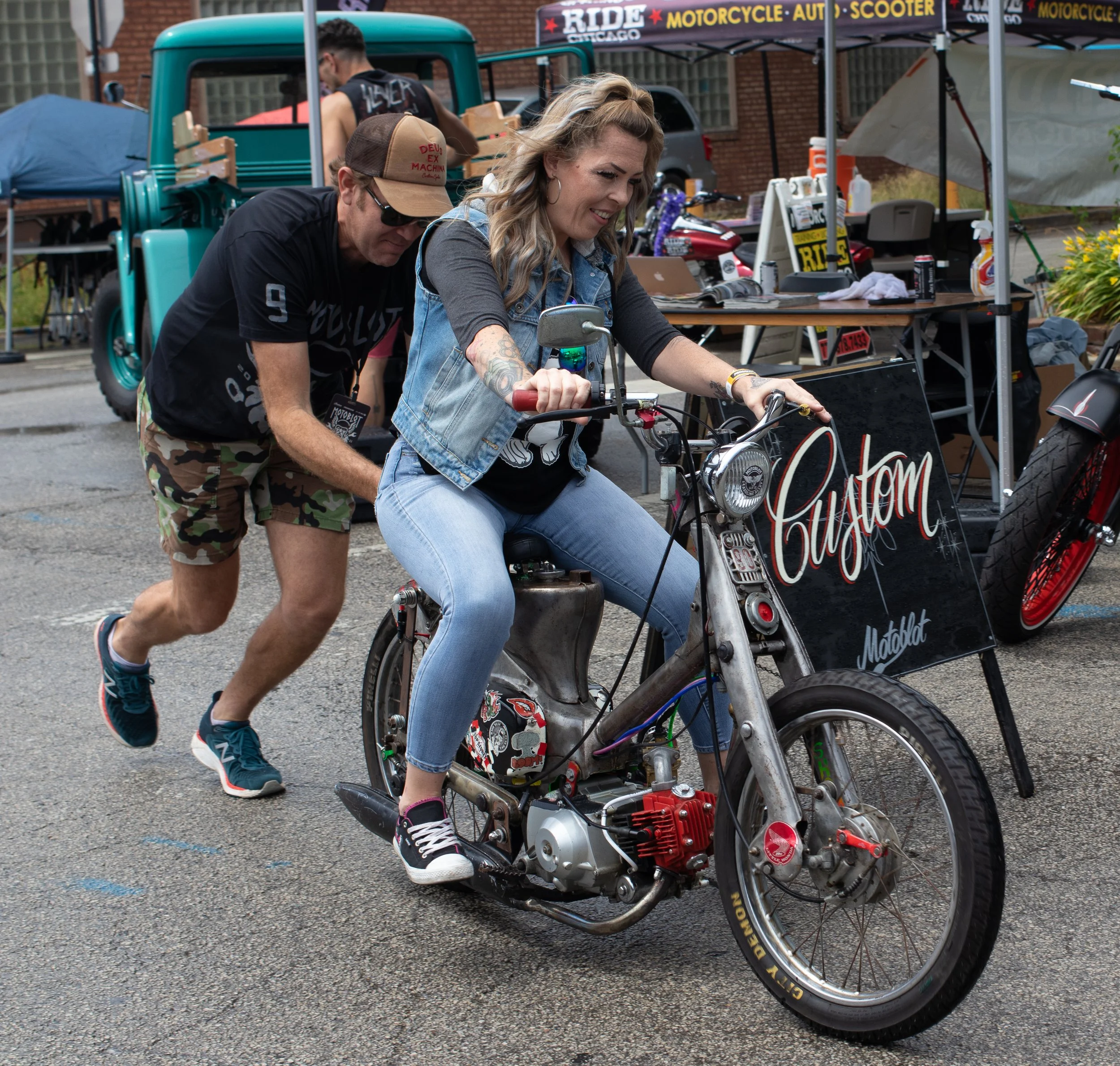 A woman is sitting on a motorcycle at an outdoor market, with a man leaning over her. The market has tents and booths, and a teal vintage truck is visible in the background. The woman is smiling and wearing a denim vest and jeans, while the man is we