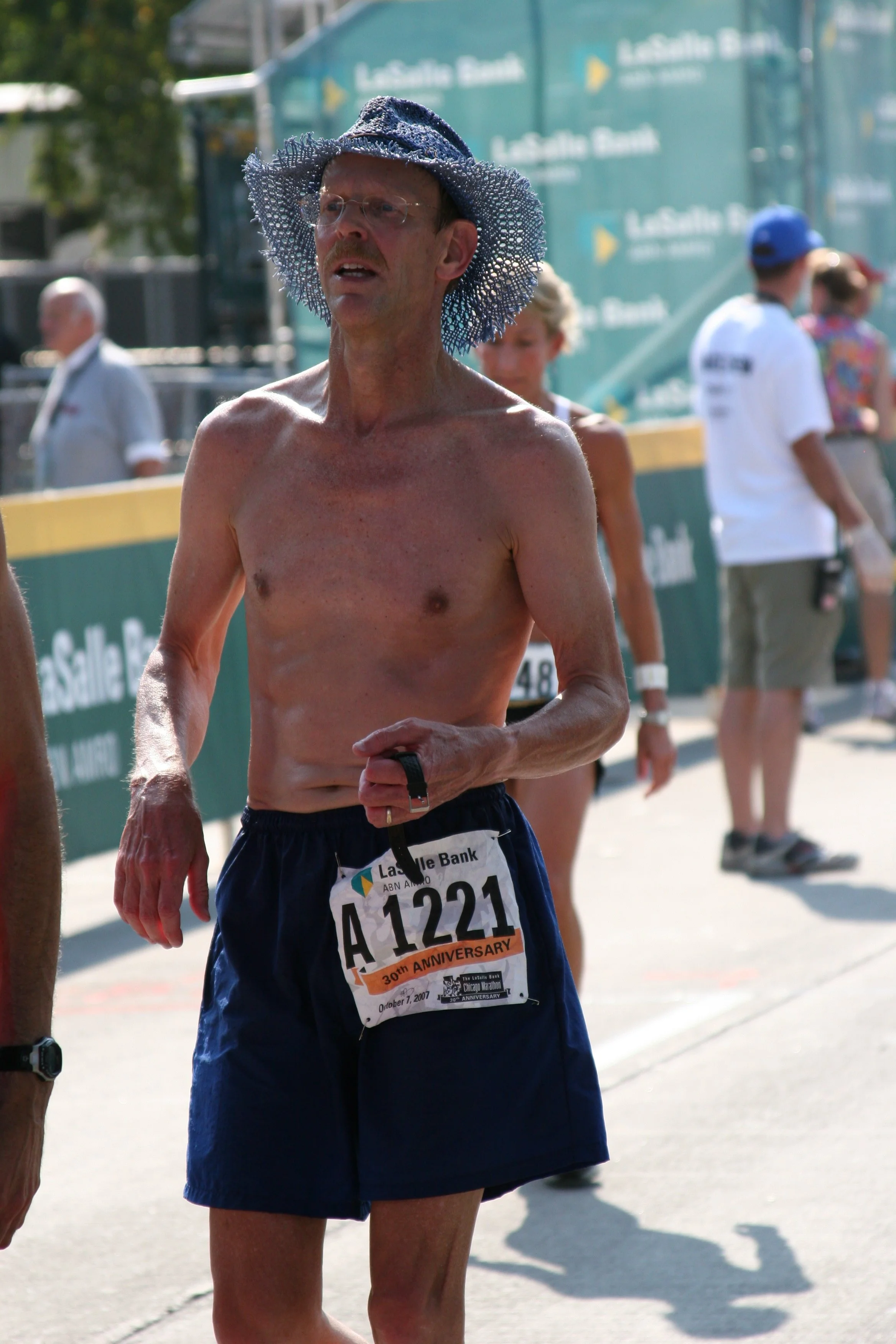 A shirtless man wearing glasses and a wide-brimmed, netted blue hat, running in a race with a bib number A 1221, during daytime at an outdoor event, with other participants and spectators in the background.