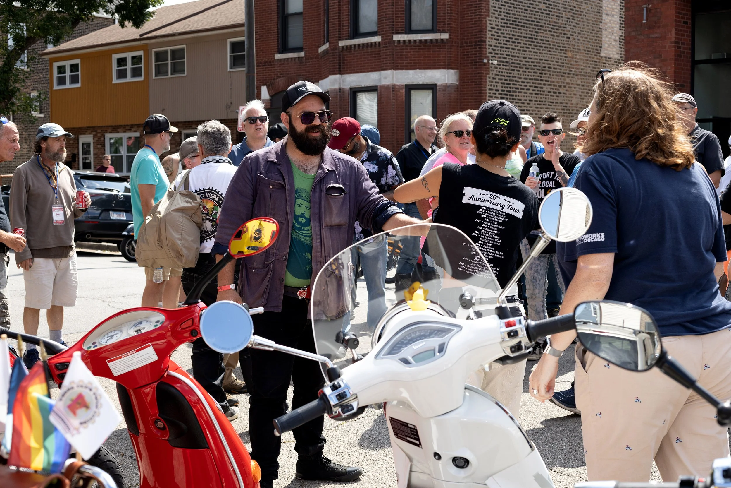 A group of people gathered outdoors around scooters and motorcycles on a sunny day, with brick and beige buildings in the background.
