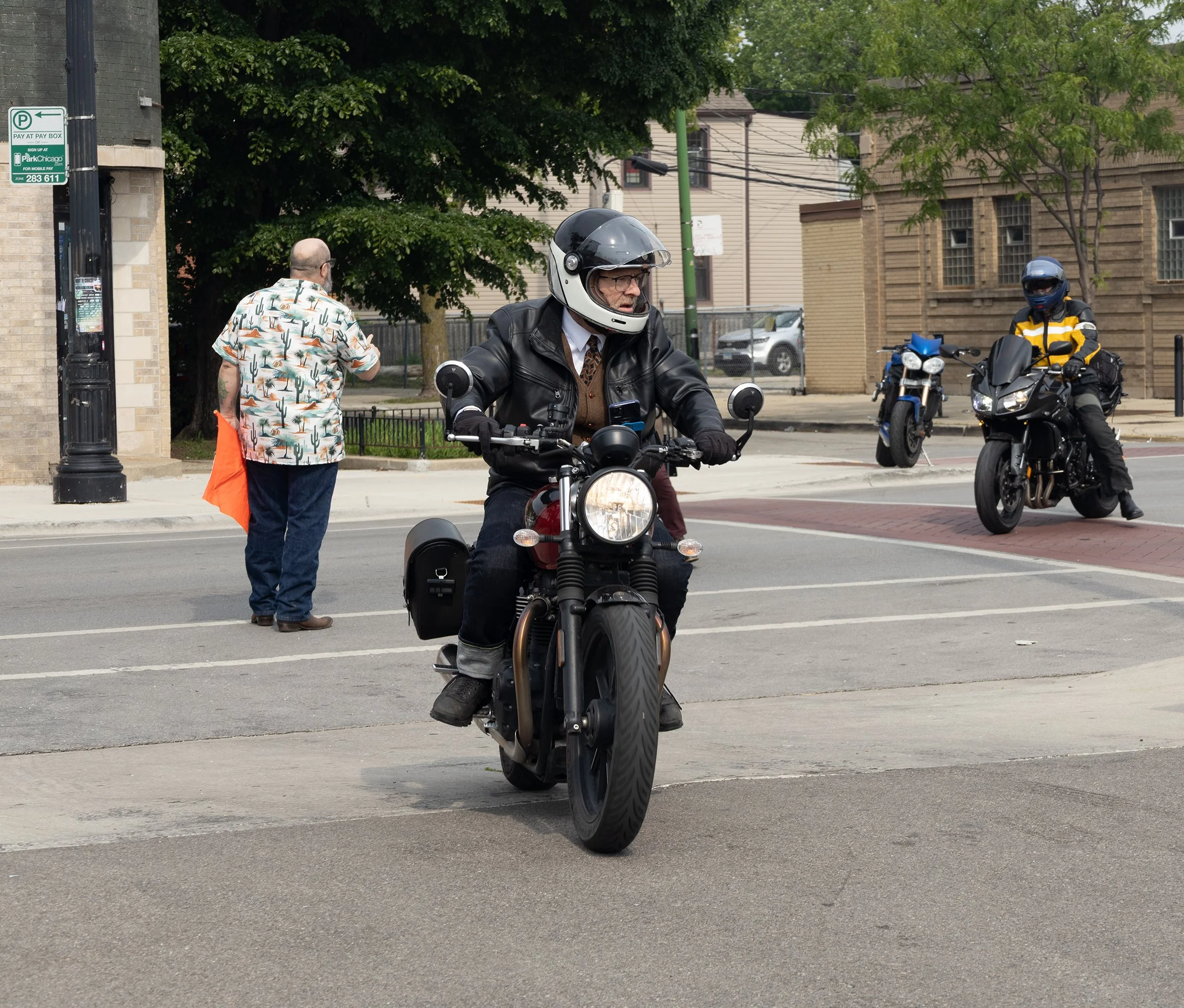 A man riding a motorcycle wears a helmet and glasses, and a man in a Hawaiian shirt and orange bag stands nearby, with two other motorcyclists in the background at an urban intersection under trees and buildings.