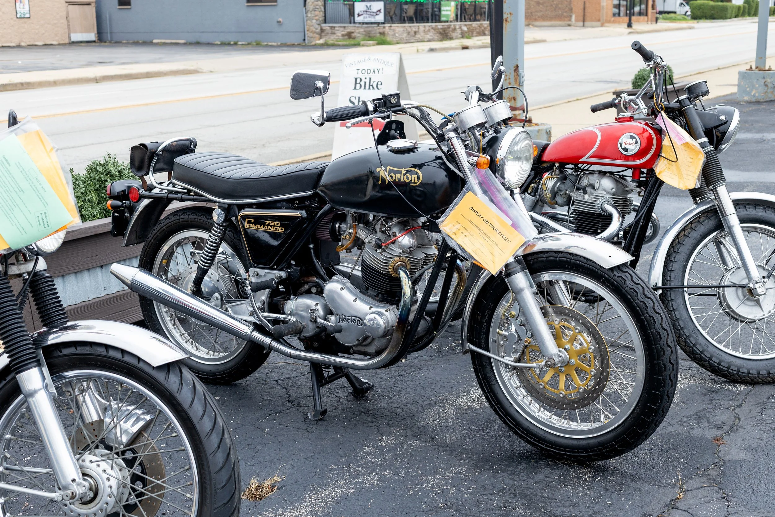 Two vintage Norton motorcycles parked outside a shop, with signage and other bikes in the background.