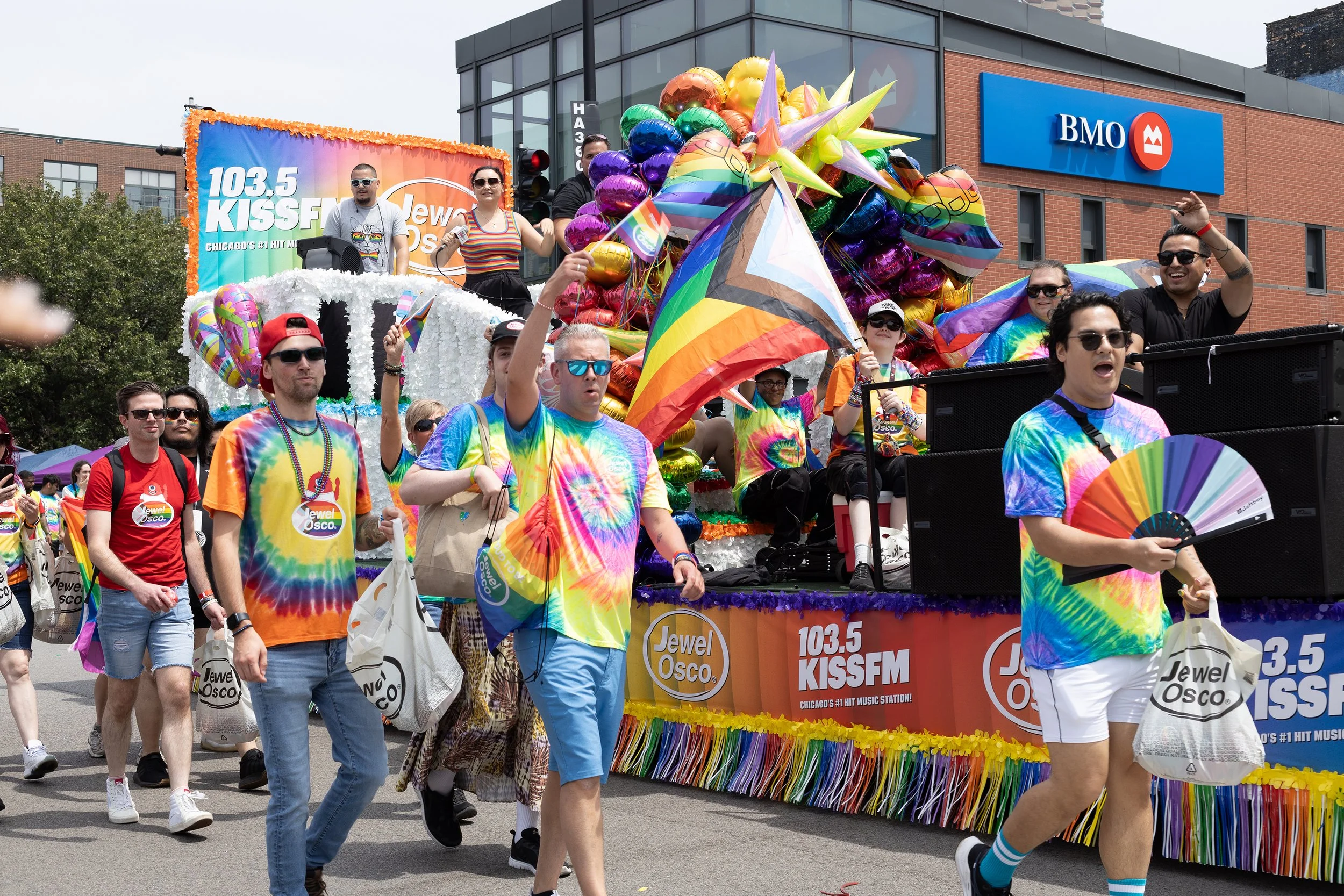 A colorful parade float decorated with rainbow balloons and banners for Jewel-Osco and 103.5 KISS FM, with people walking beside and on the float during a pride parade in an urban area.