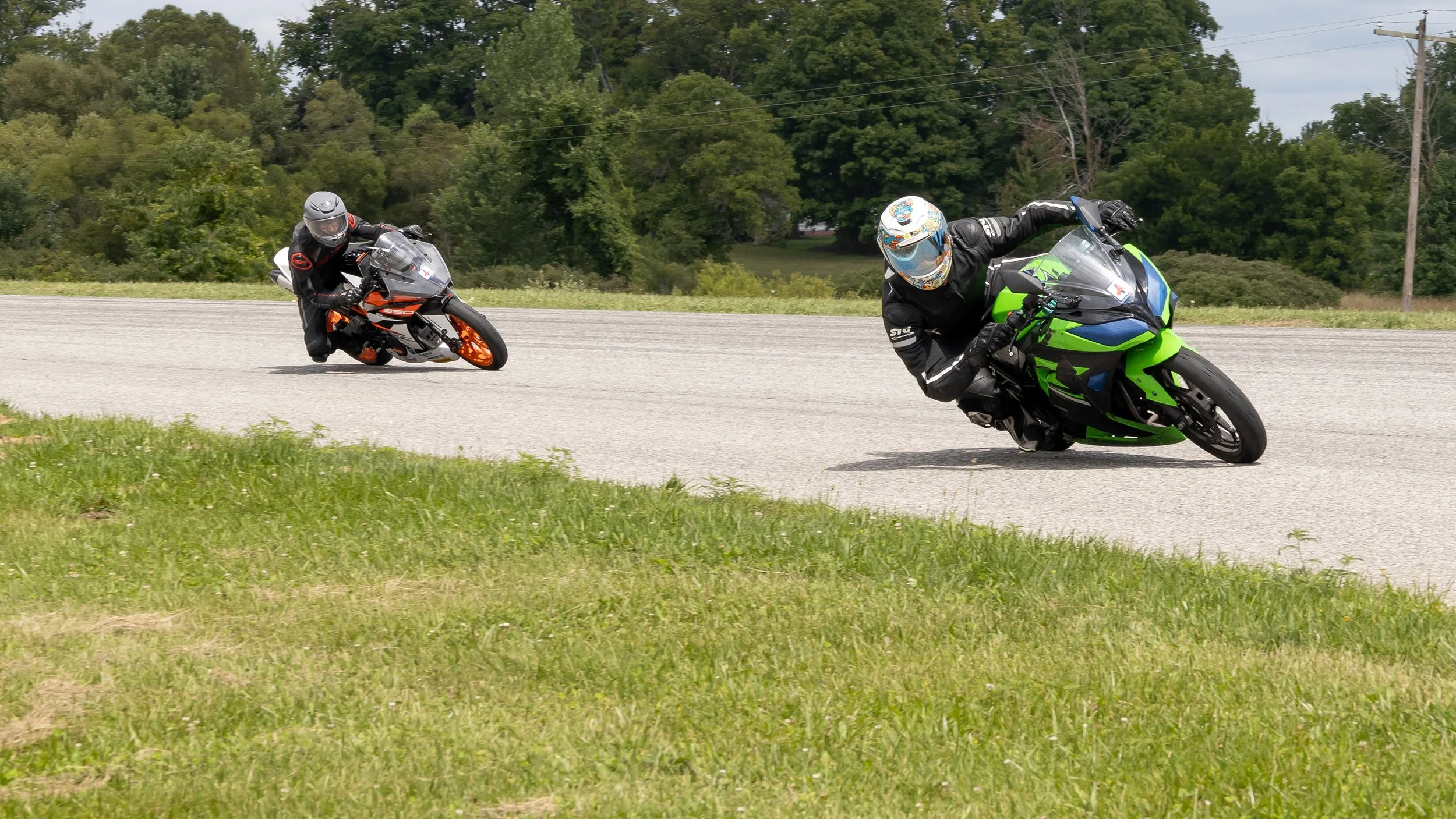 Two motorcyclists leaning sharply into a turn on a paved road, one on a black and neon green bike and the other on a black and orange bike, with green trees and power lines in the background.