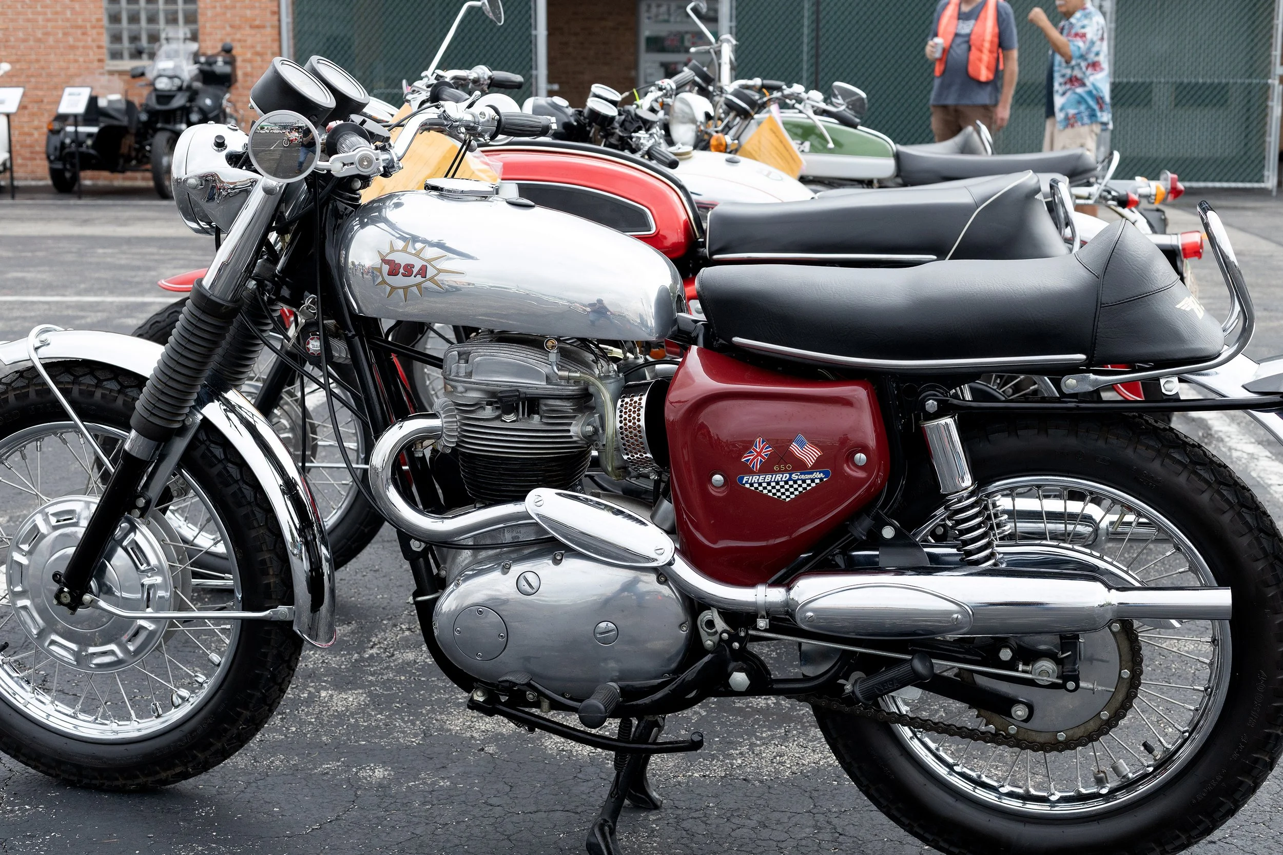 Multiple vintage motorcycles parked in a row outdoors, with people standing and talking in the background.