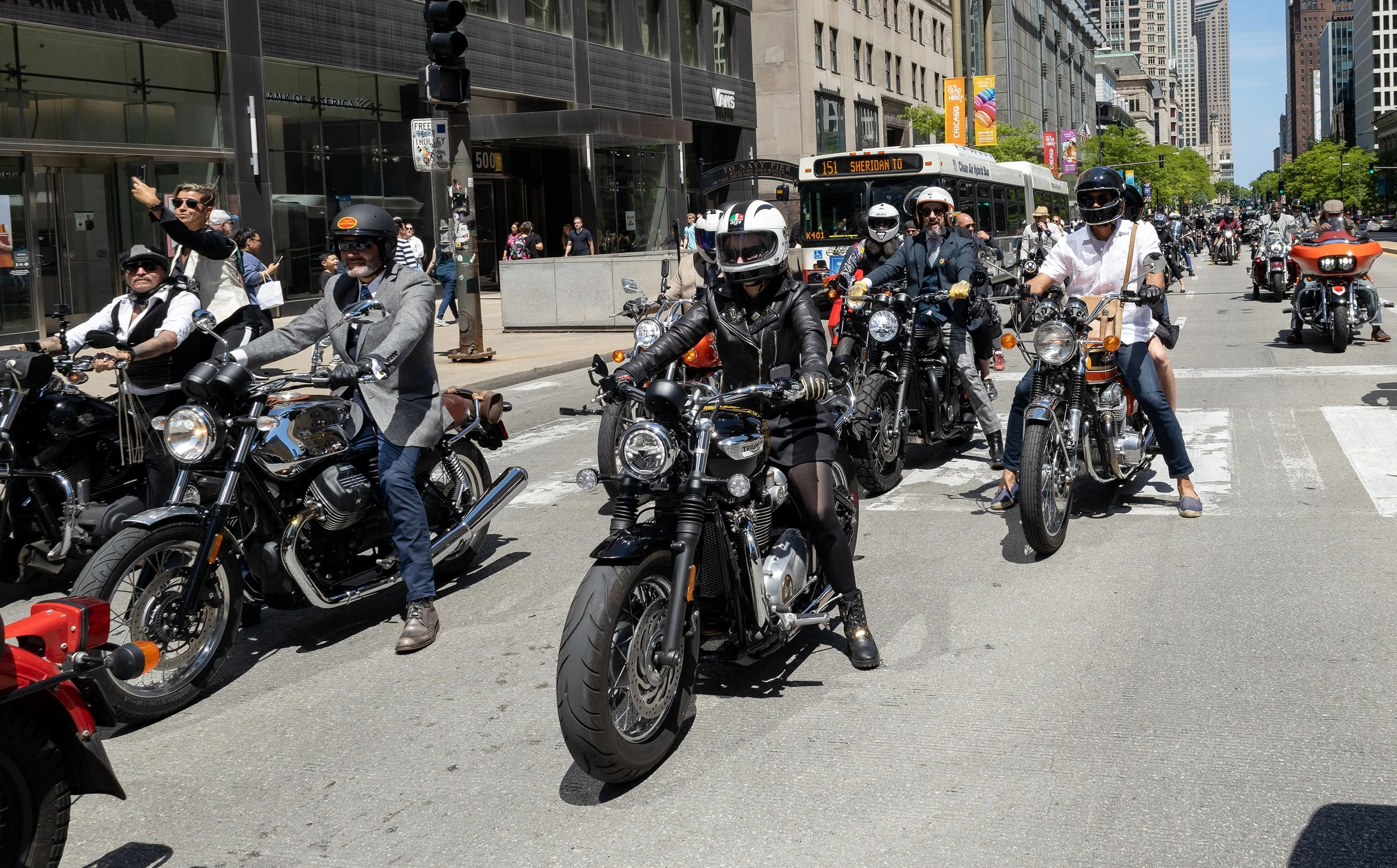 A packed city street with numerous motorcycles and riders waiting at an intersection, surrounded by tall buildings and a city bus in the background on a sunny day.