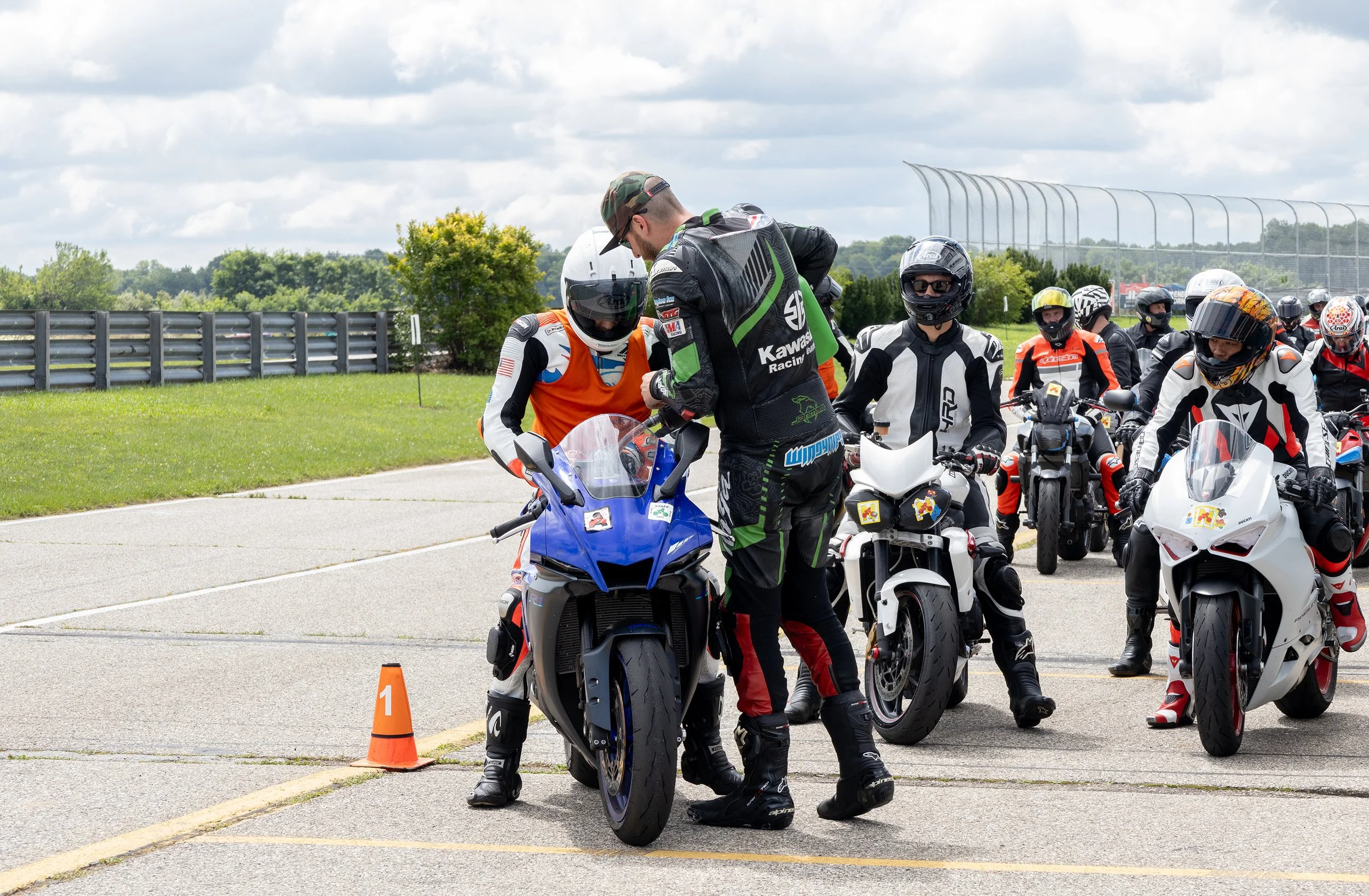 Group of motorcyclists gathered outdoors on a paved surface, some wearing helmets and racing gear, with orange cones and a numbered marker nearby, under a cloudy sky.