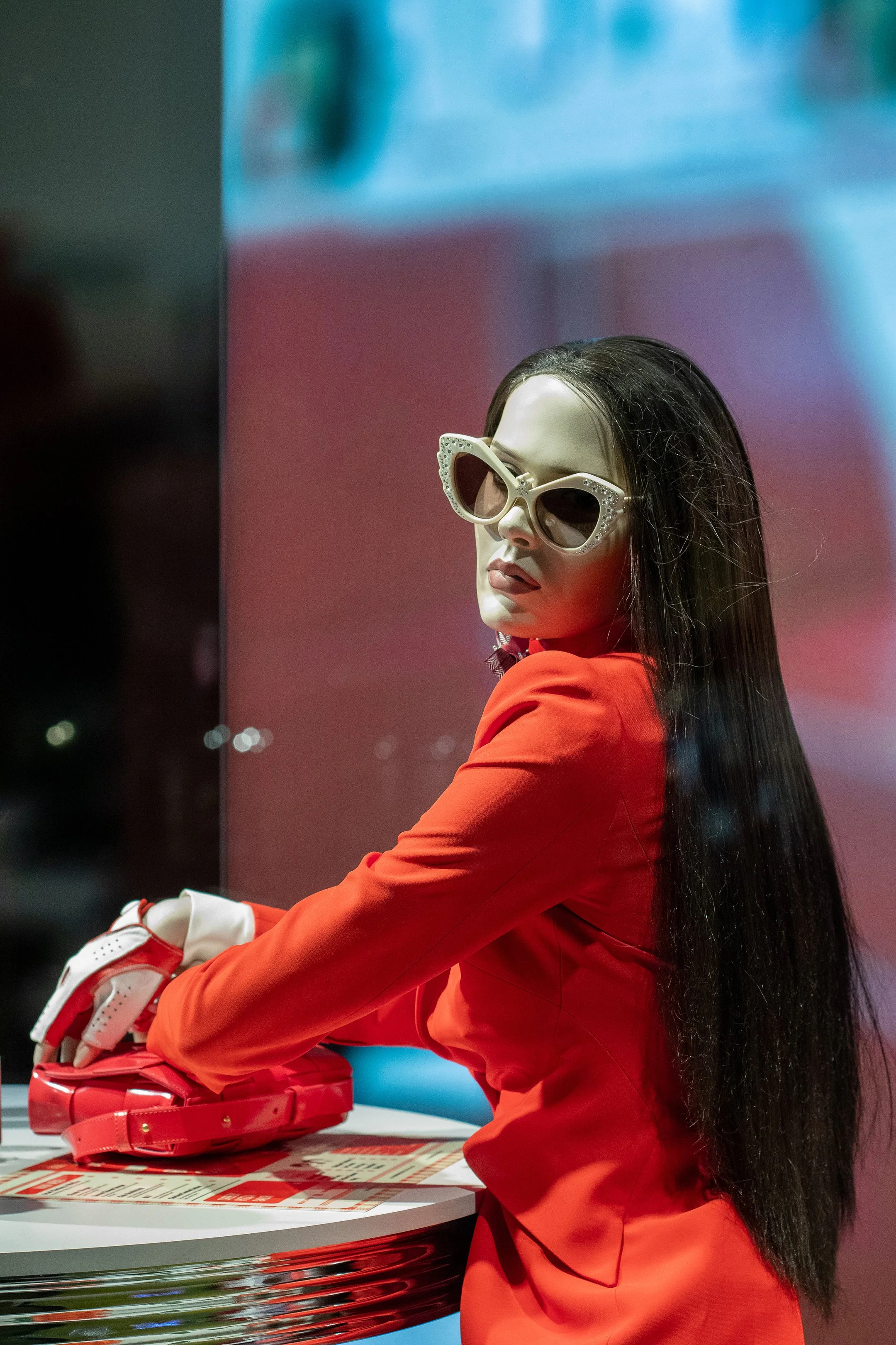 A woman with long dark hair wearing large, fashion sunglasses and a red outfit, sitting at a table with a red purse and gloves, in front of a colorful, blurred background.