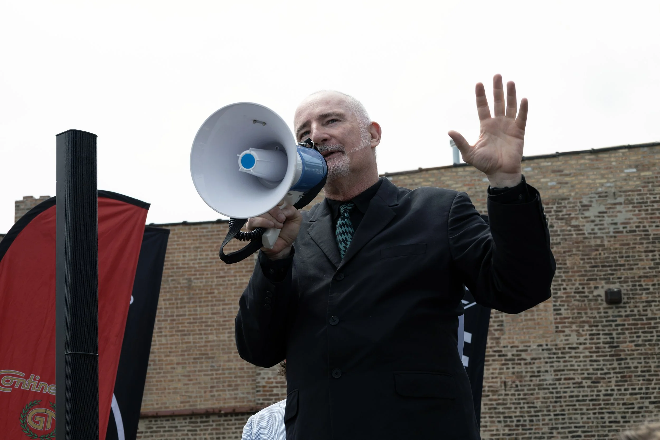 A man with a bald head and gray beard speaking into a blue and white megaphone while raising his left hand. He is wearing a black suit and a stylish tie. The background features a brick wall and flags, indicating an outdoor event.