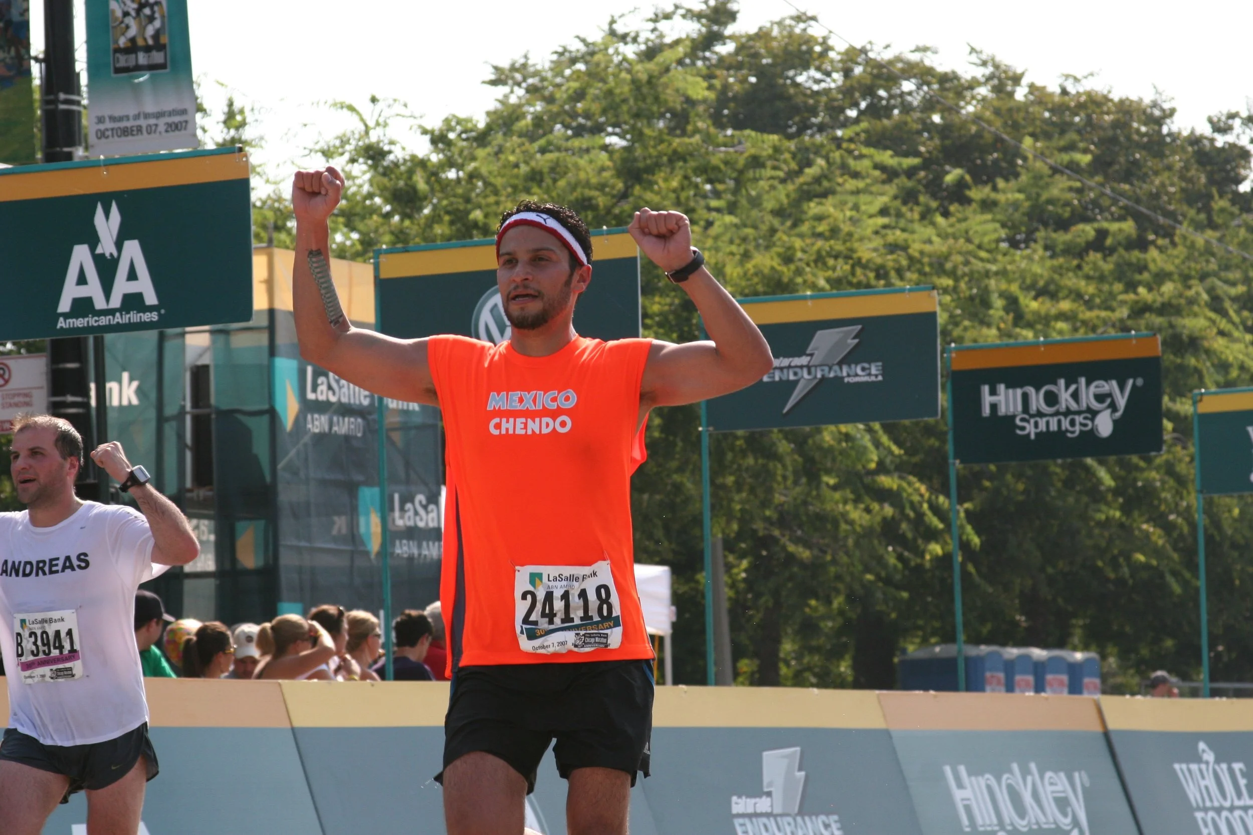 A male marathon runner in a bright orange shirt with 'Mexico Chendo' printed on it, raising his arms in celebration or victory as he crosses the finish line in a race. He is wearing a red and white headband, black shorts, and a wristwatch, with a cro