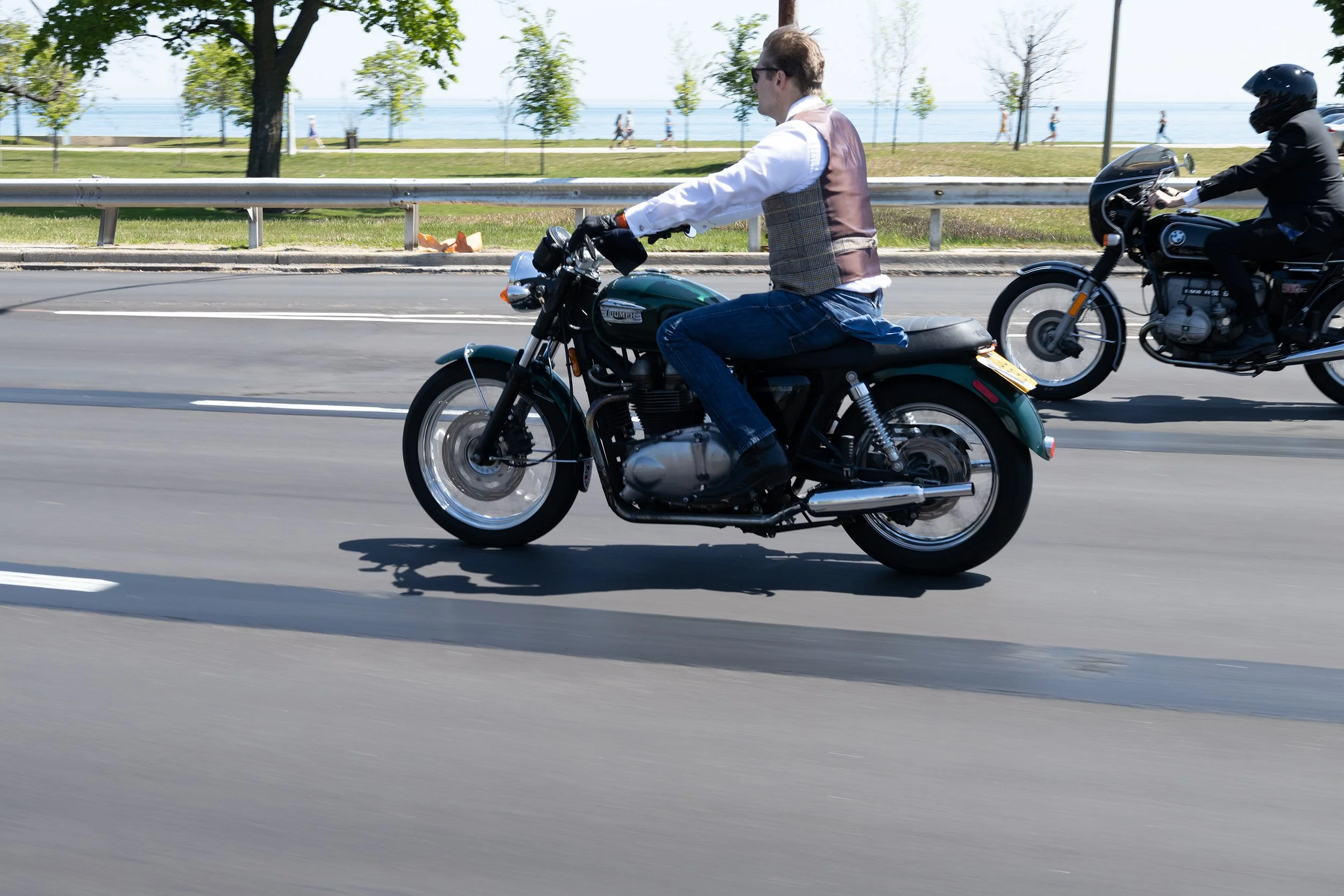 A man riding a Triumph motorcycle and a person on a BMW motorcycle on a city street near a park and water.