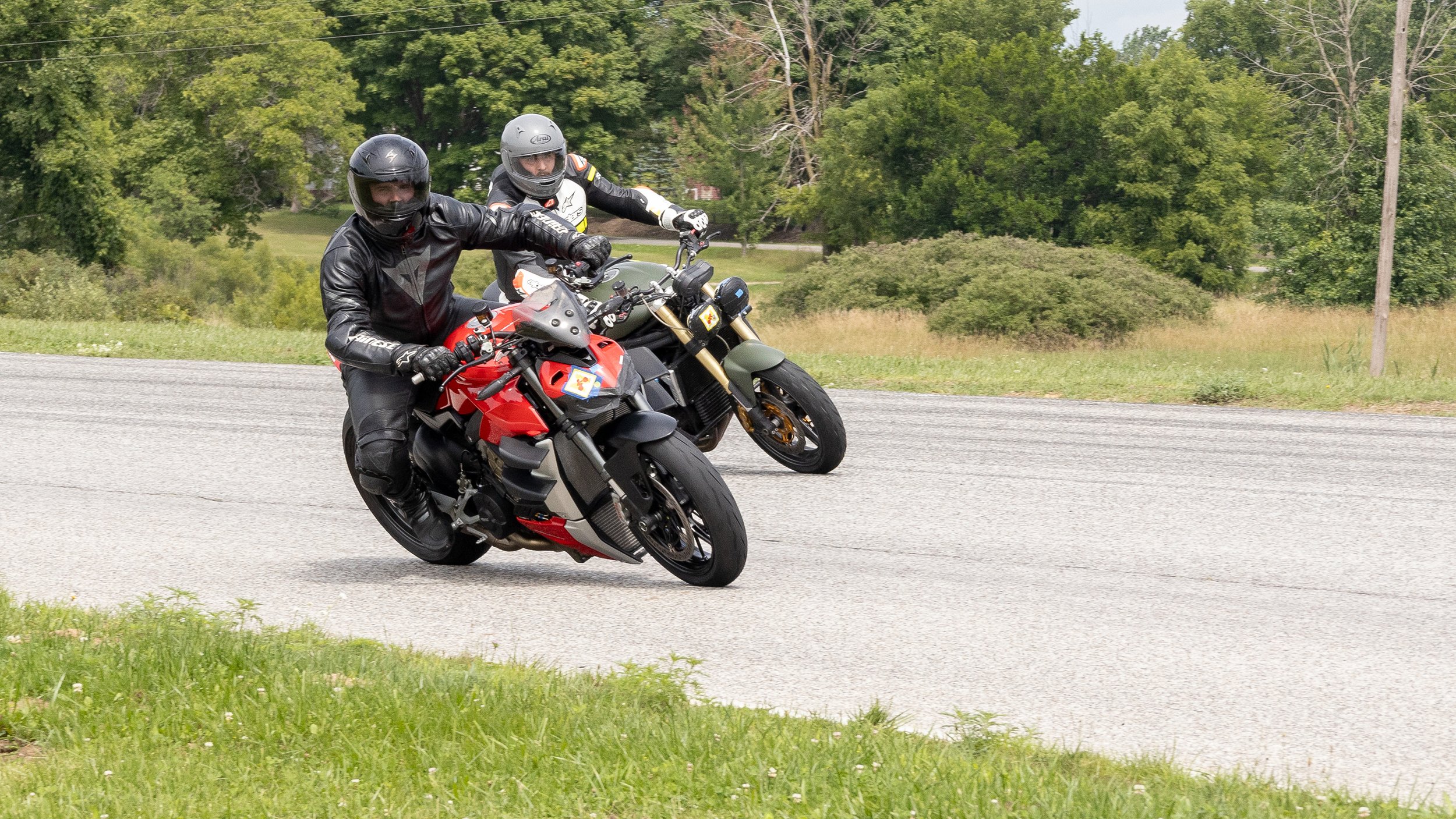 Two motorcyclists riding on a rural road surrounded by green trees and grass, wearing black and gray helmets and riding sport bikes, one red and one black, with a cloudy sky overhead.