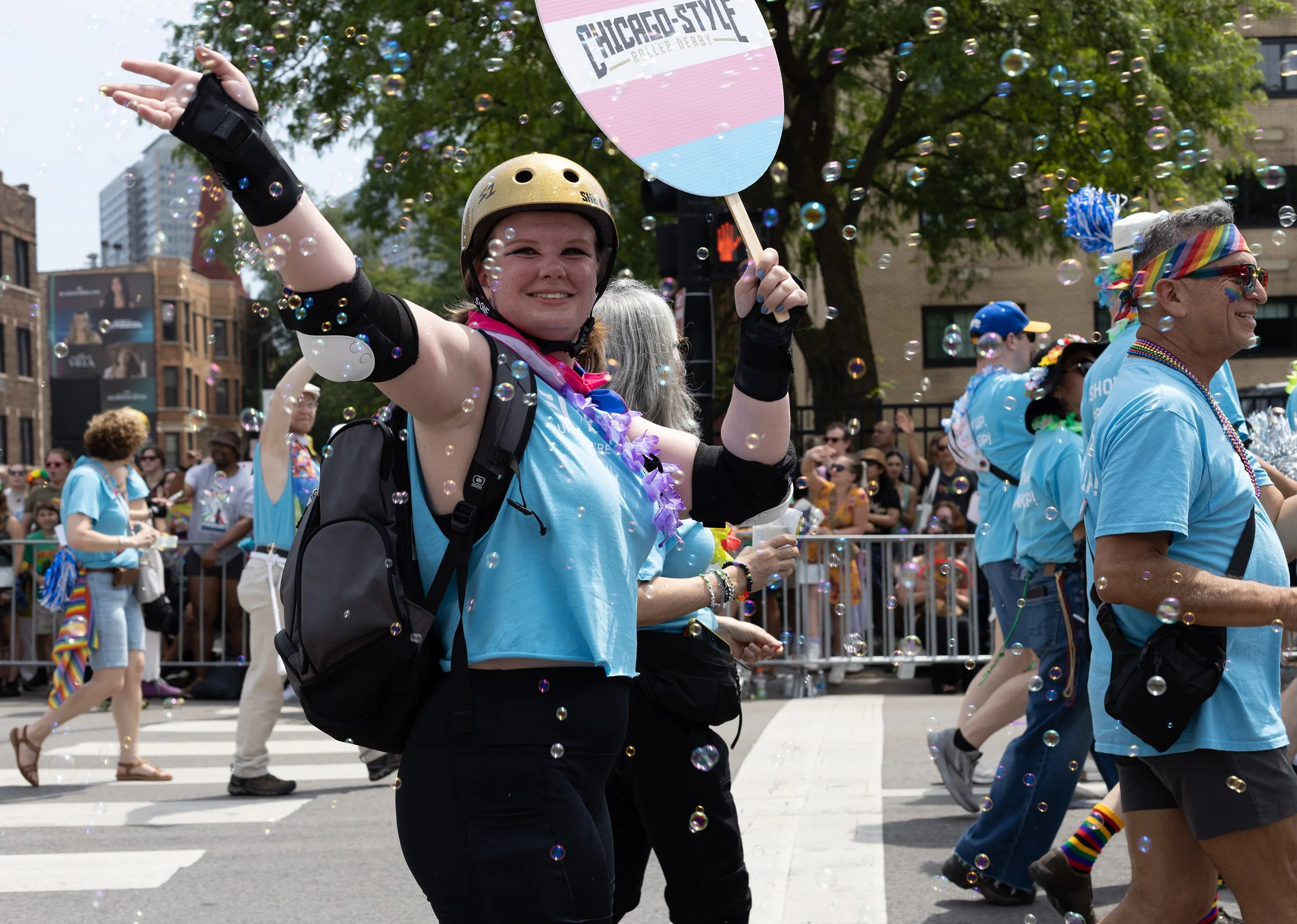 People celebrating in a street parade during Pride with rainbow accessories, bubbles, and colorful outfits, one person holding a sign that reads "Chicago Style Roller Derby".