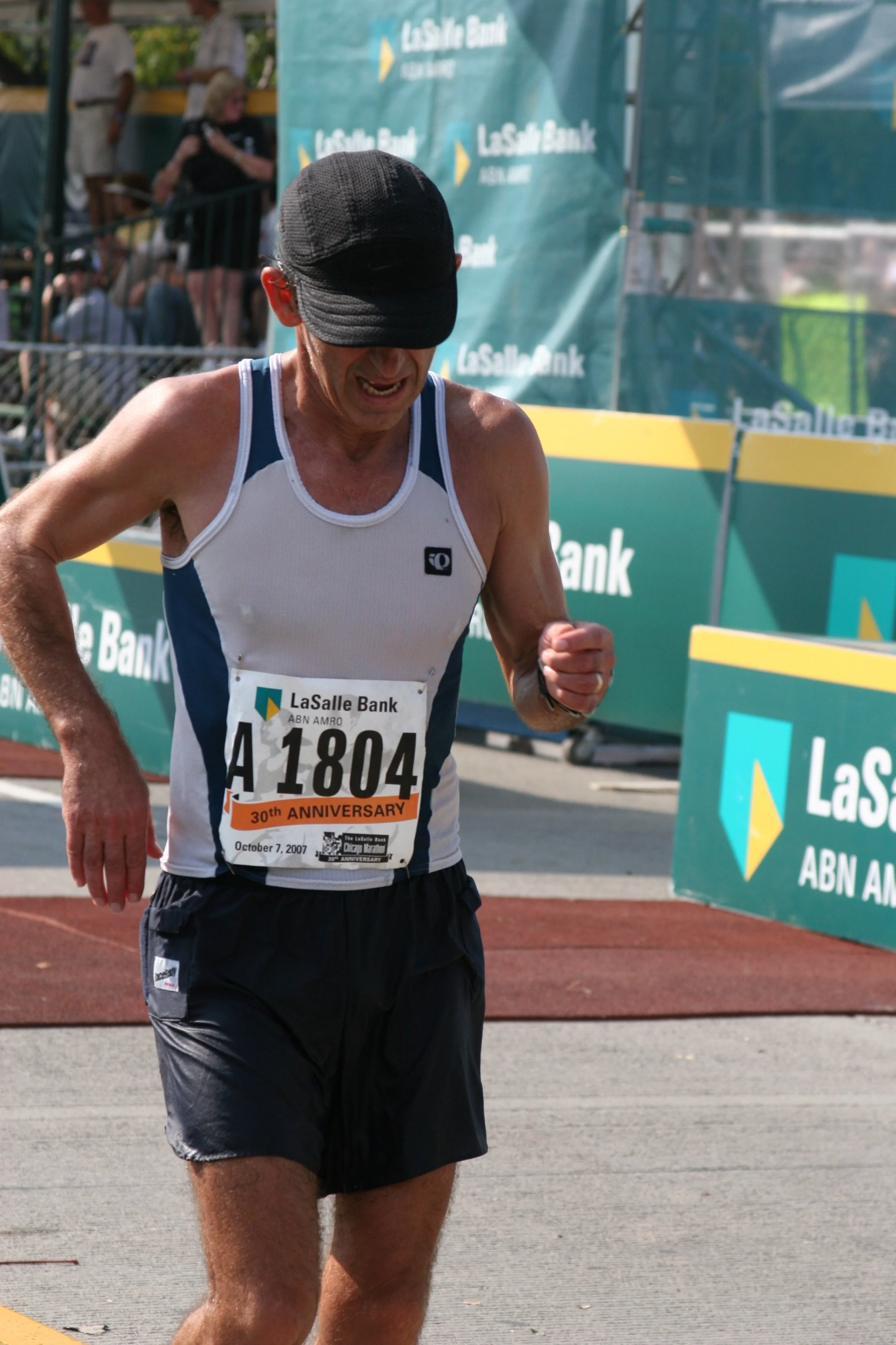 A male marathon runner wearing a black cap and a white and blue athletic shirt with a bib numbered 41804, visibly sweating and tired, crossing a finish line at a marathon event with 