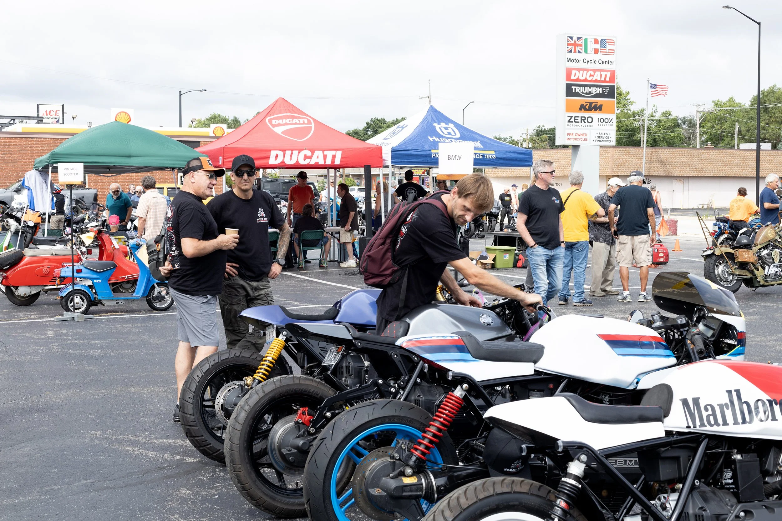 A motorcycle event in a parking lot with people gathered around motorcycles, tents with brand logos, and a sign listing various motorcycle brands.