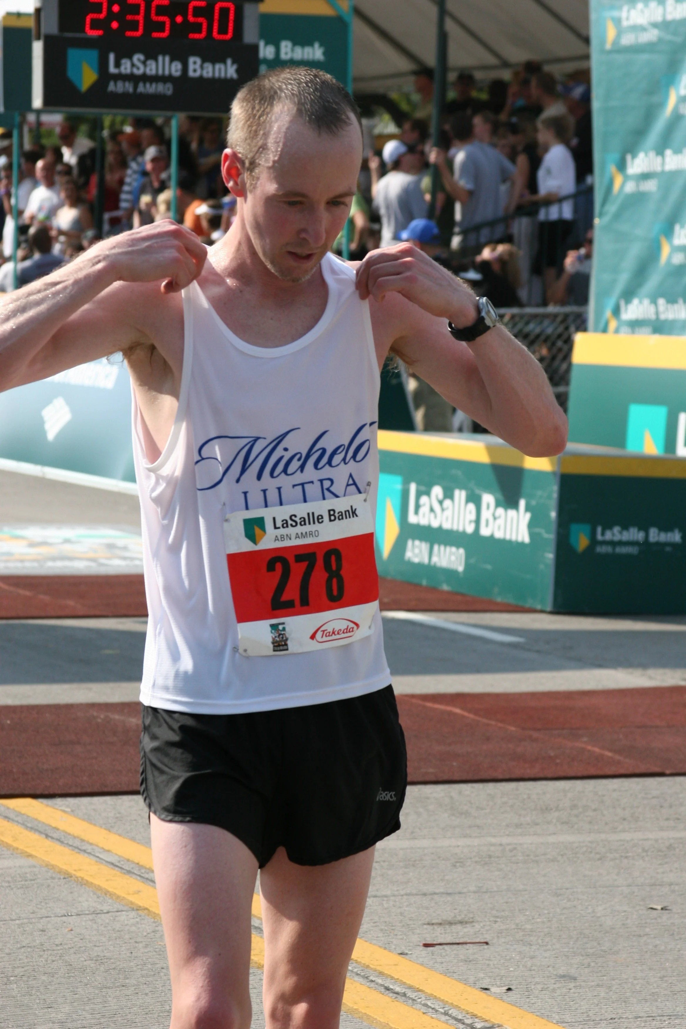 A male marathon runner wearing a white tank top with 'Michelo Ultra' written on it, black shorts, and a watch on his left wrist, adjusting his shirt after crossing the finish line at a race event, with a digital timer displaying 2:35:50 and a crowd o