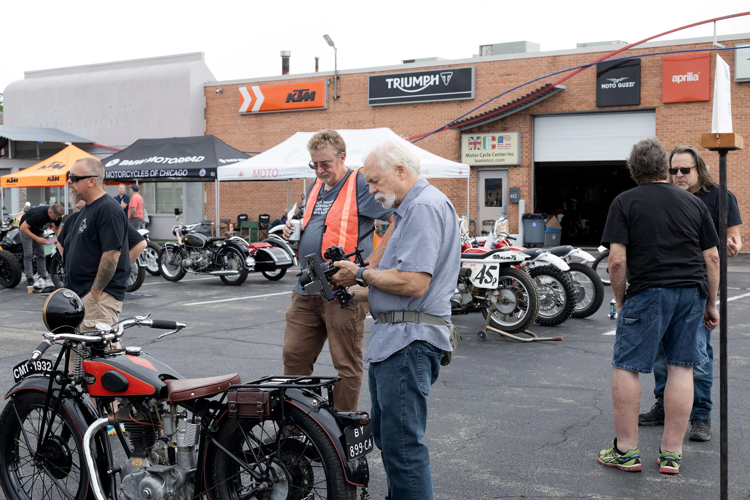 A gathering of motorcycle enthusiasts outdoors, with vintage and modern bikes on display, some people examining the motorcycles, and branded tents and signs for KTM, Triumph, Moto Guzzi, and Aprilia in the background.