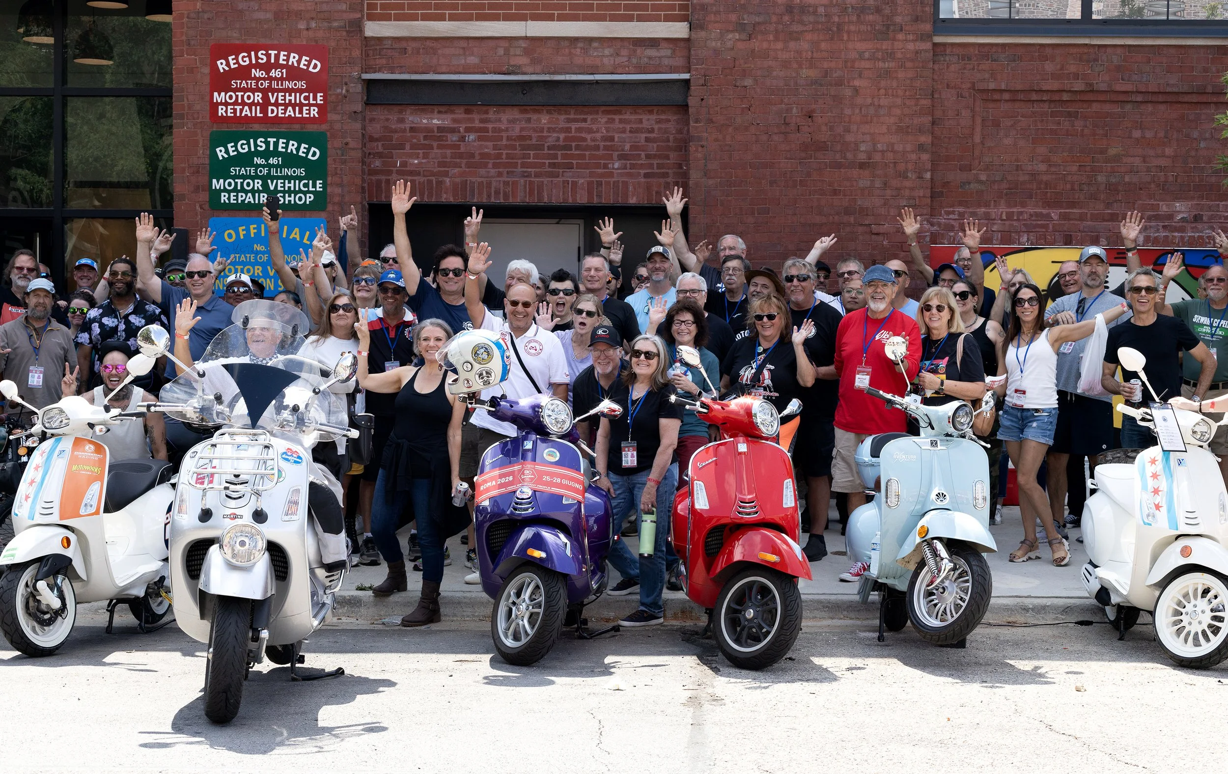 A group of people gathered outside a brick building, raising their hands and smiling, with scooters parked in front of them.