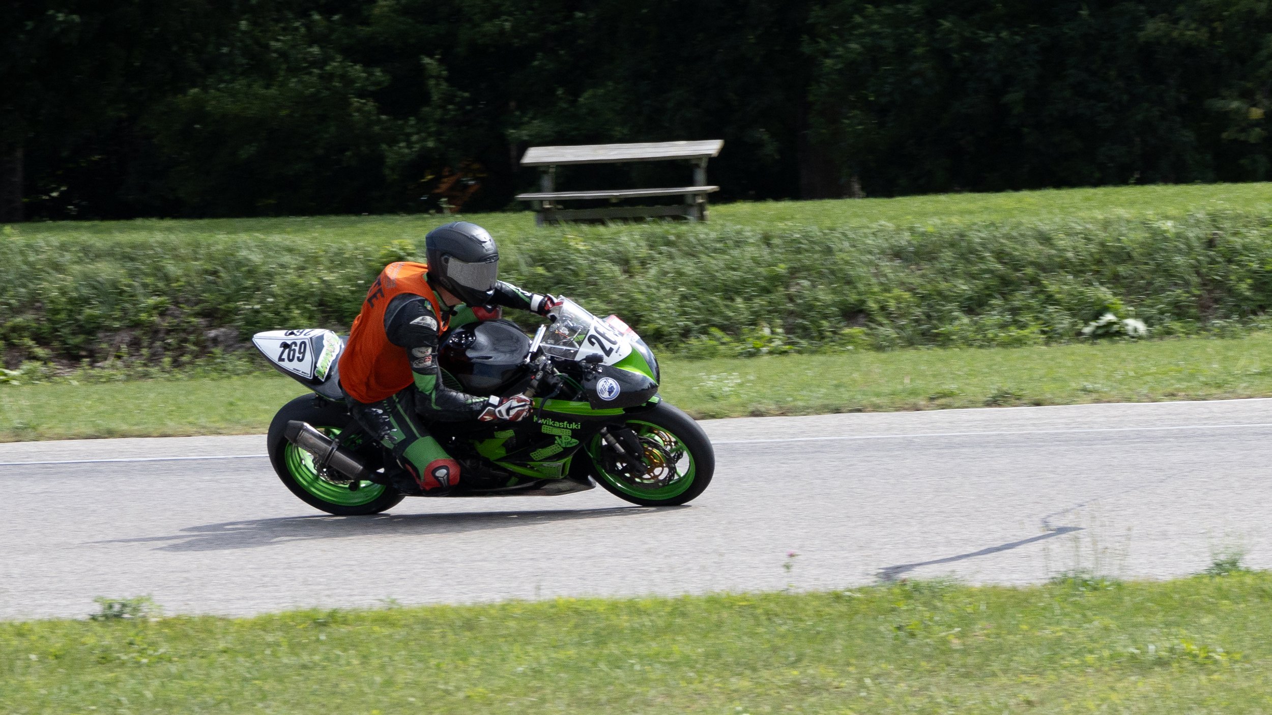 A motorcyclist wearing a black helmet, black, green, red, and white racing gear, and an orange vest riding a green and black sports bike on a paved road with green grass and trees in the background.