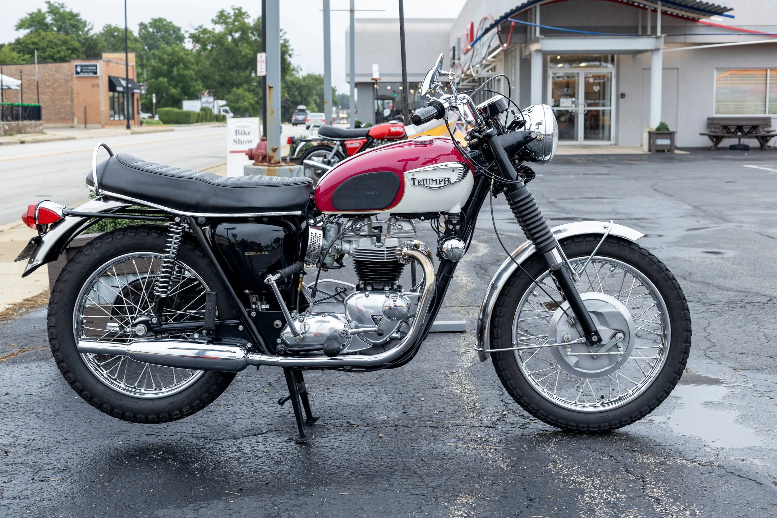 A red and white vintage Triumph motorcycle parked outside a building on a wet pavement.