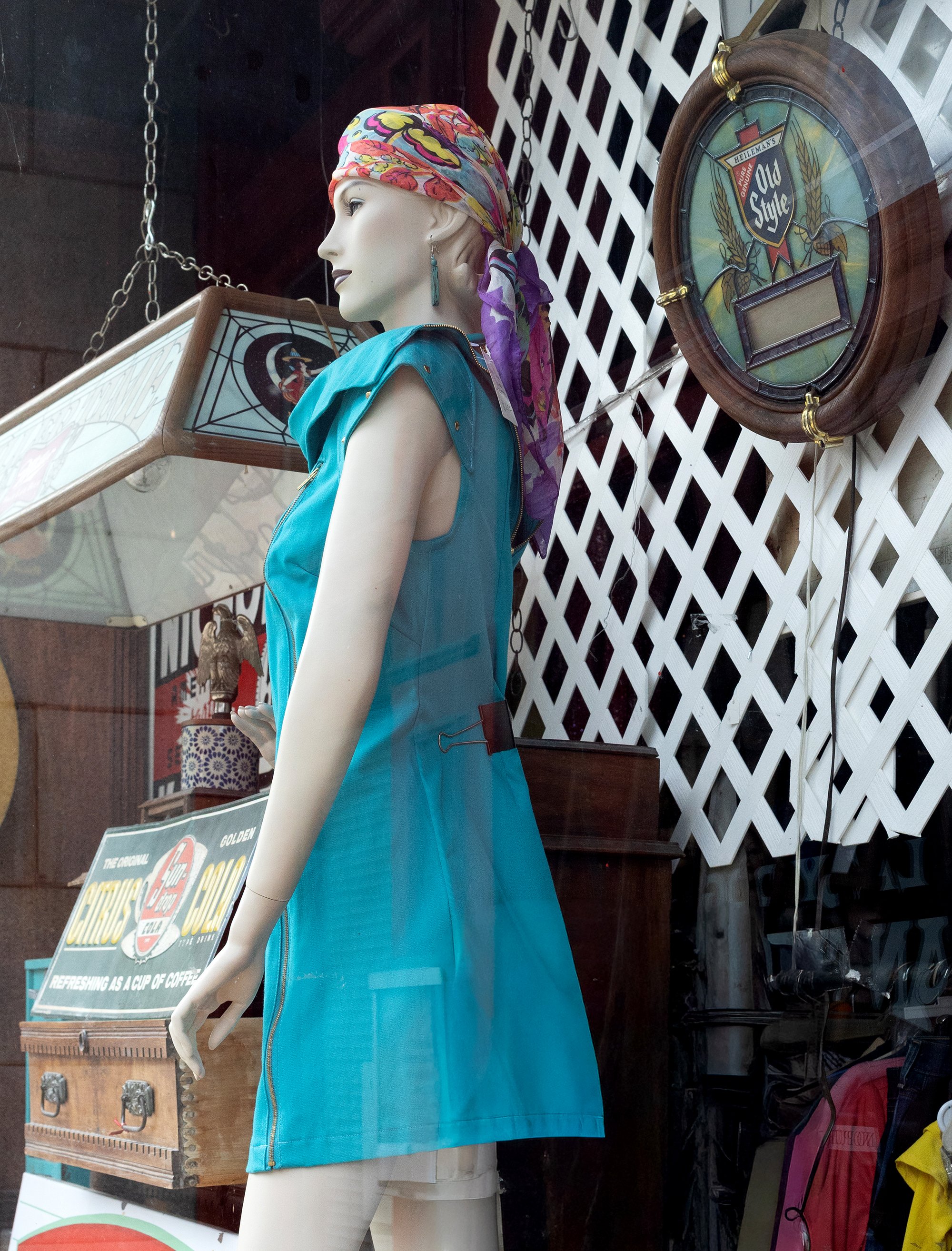 A mannequin wearing a colorful headscarf and a sleeveless blue jacket in a storefront display, with vintage signs and decorative items in the background.