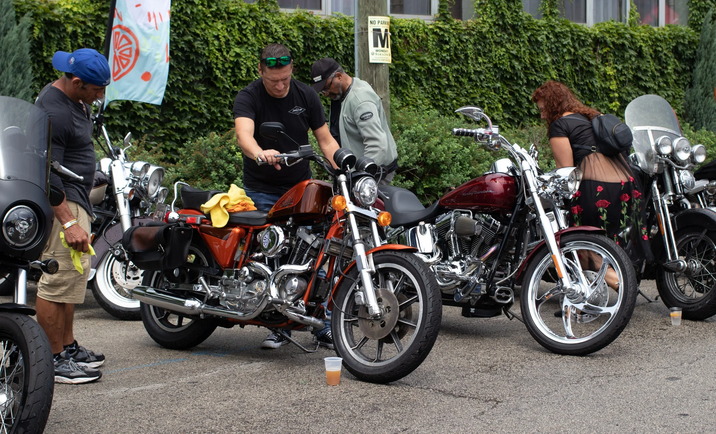 Group of people gathered around parked motorcycles on a city street with a green ivy-covered wall in the background, some people inspecting the bikes, some reading. One drinks from a cup on the ground.