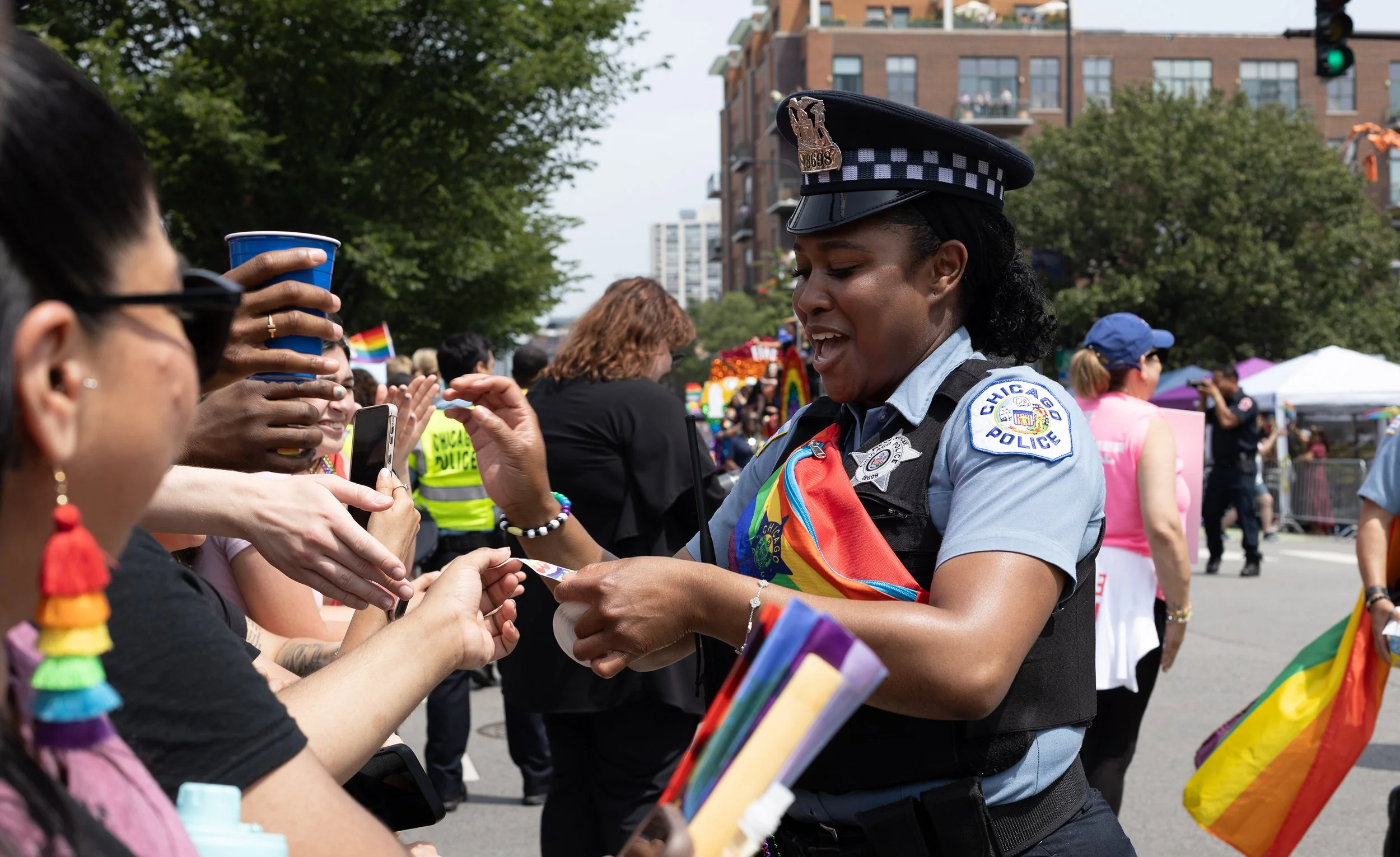 A police officer, dressed in uniform with a Chicago Police badge, is at a pride event interacting with a crowd. The officer is wearing a rainbow pride sash and is engaged in conversation with people holding rainbow flags and supplies. The background 