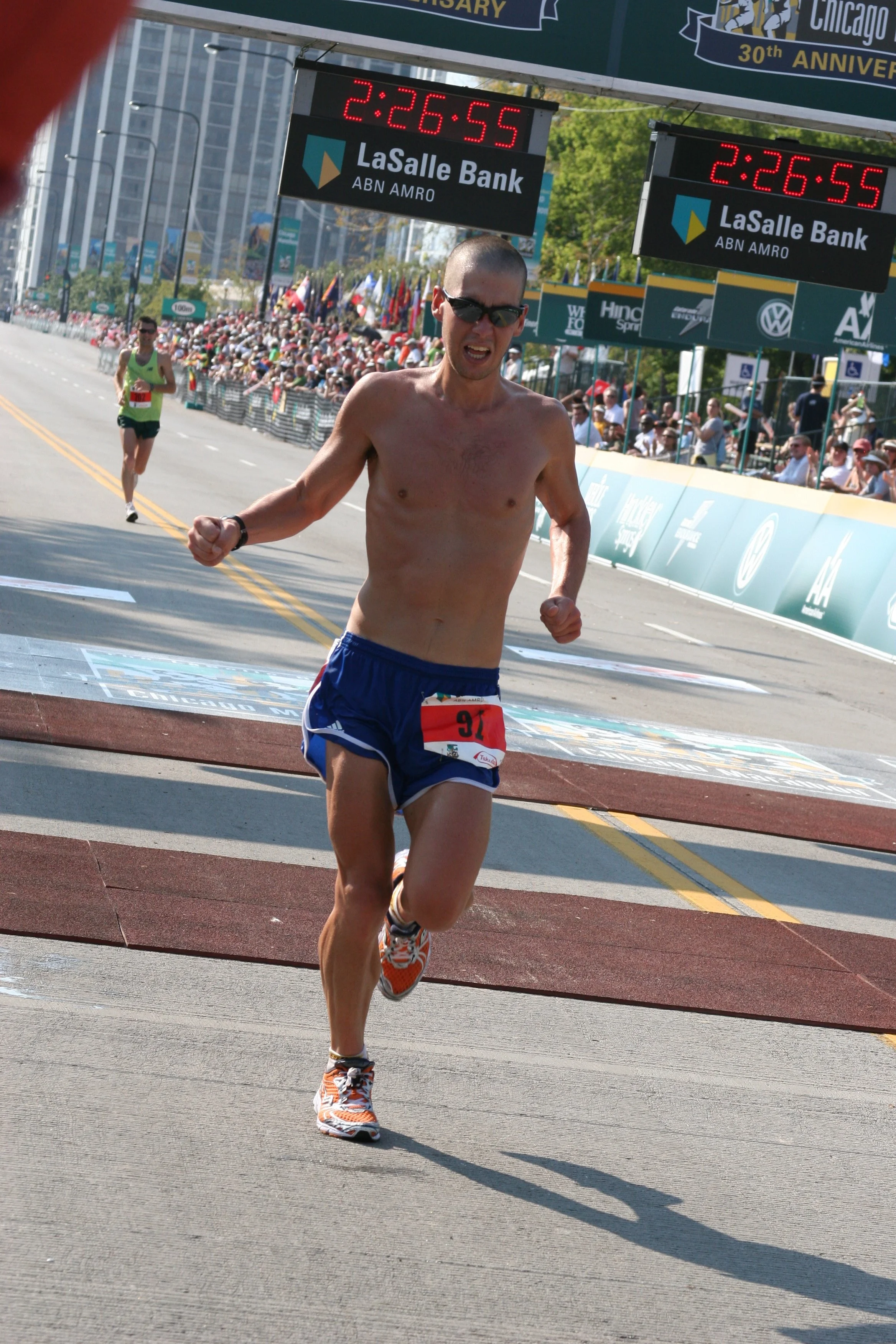 A man running shirtless in a marathon, nearing the finish line with a crowd of spectators behind barriers and a digital timer displaying 2:26:55.