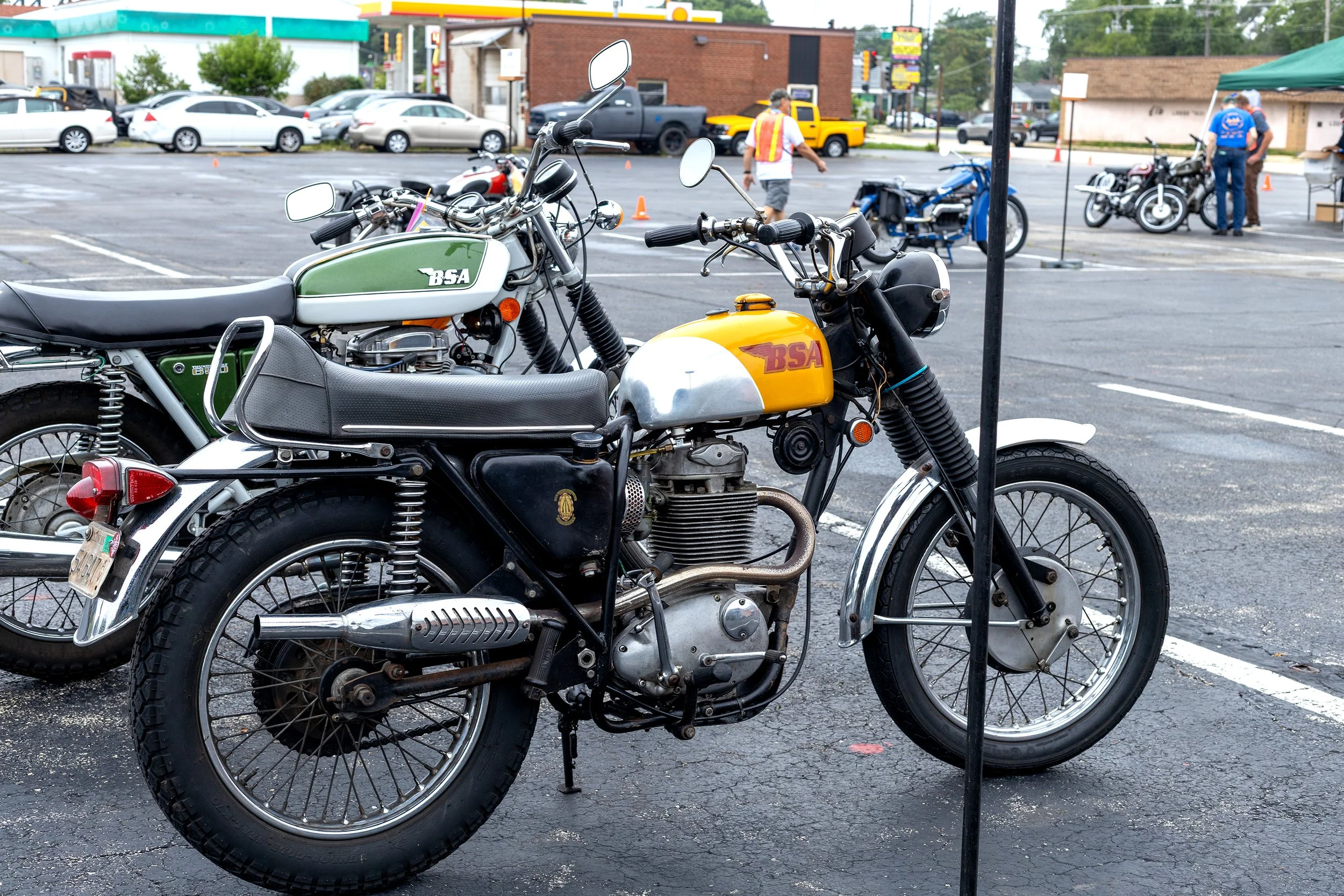 Close-up of vintage motorcycles parked in a parking lot, with a background showing people, cars, and a brick building.