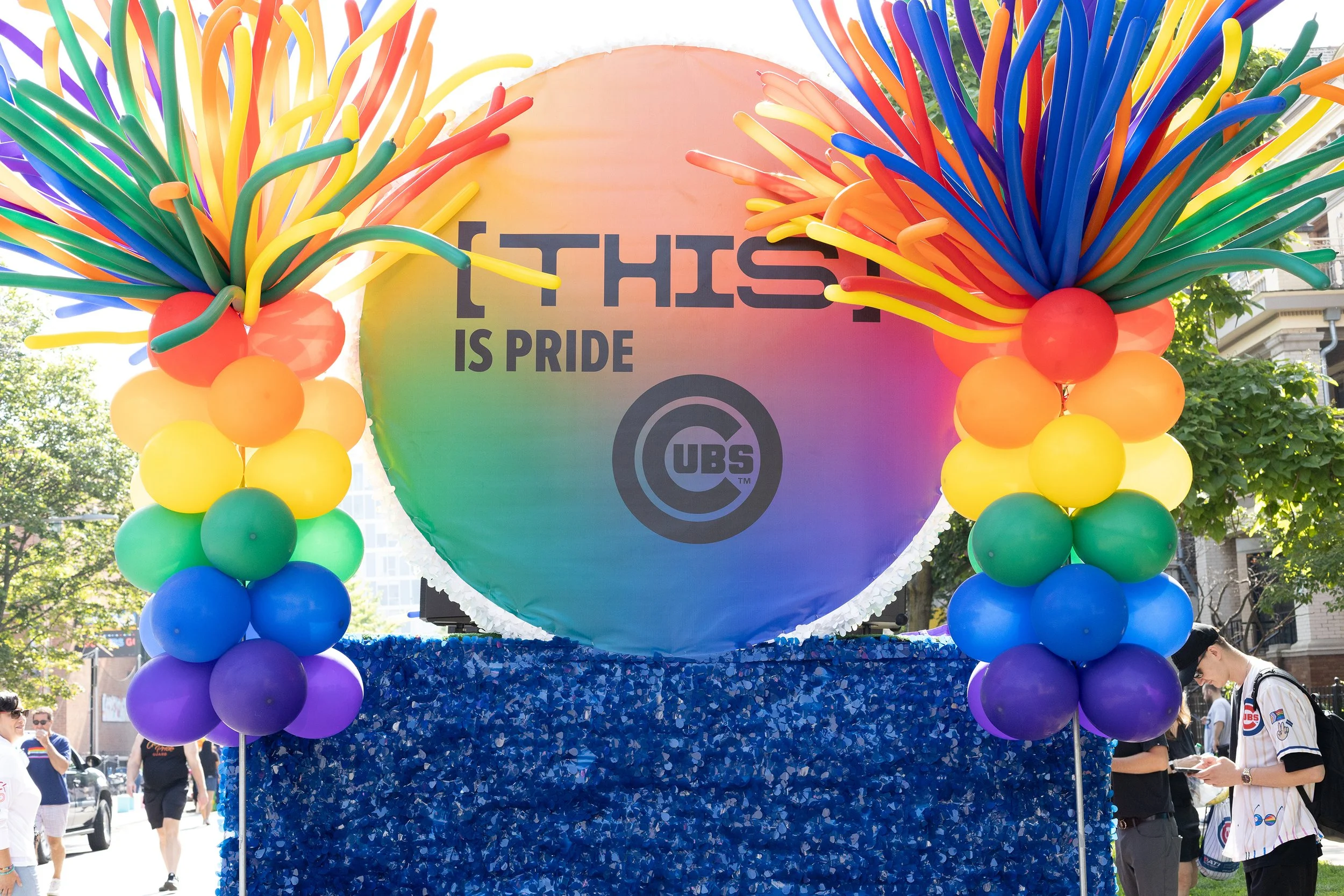 Rainbow balloon arch framing a circular sign that reads 'THIS IS PRIDE' with the logo of the Chicago Cubs baseball team underneath. The arch includes balloons in rainbow colors, and there are people in the background at an outdoor event.