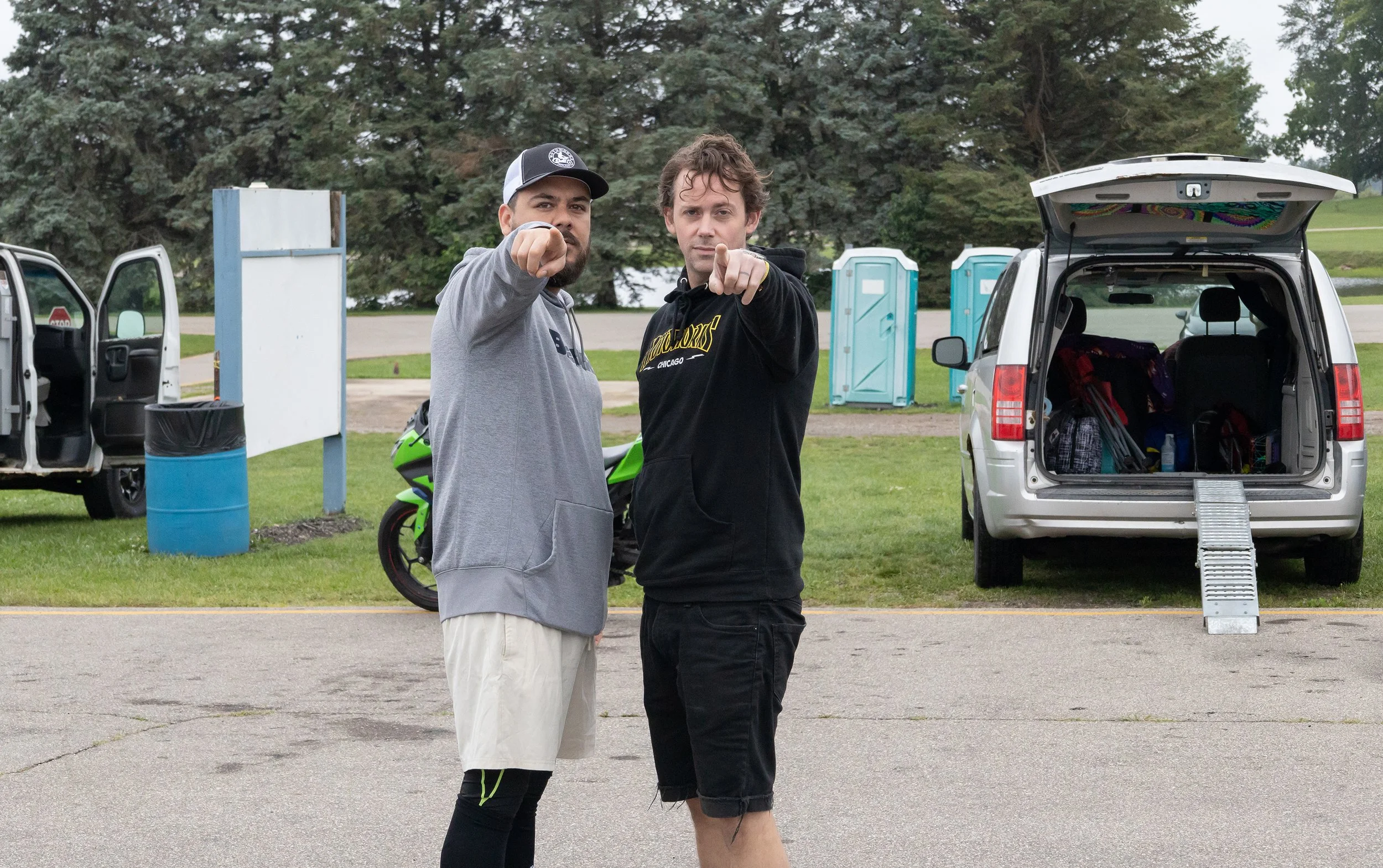 Two men pointing towards the camera stand in front of an open vehicle trunk filled with bags and supplies, with portable toilets and a green outdoor setting in the background.