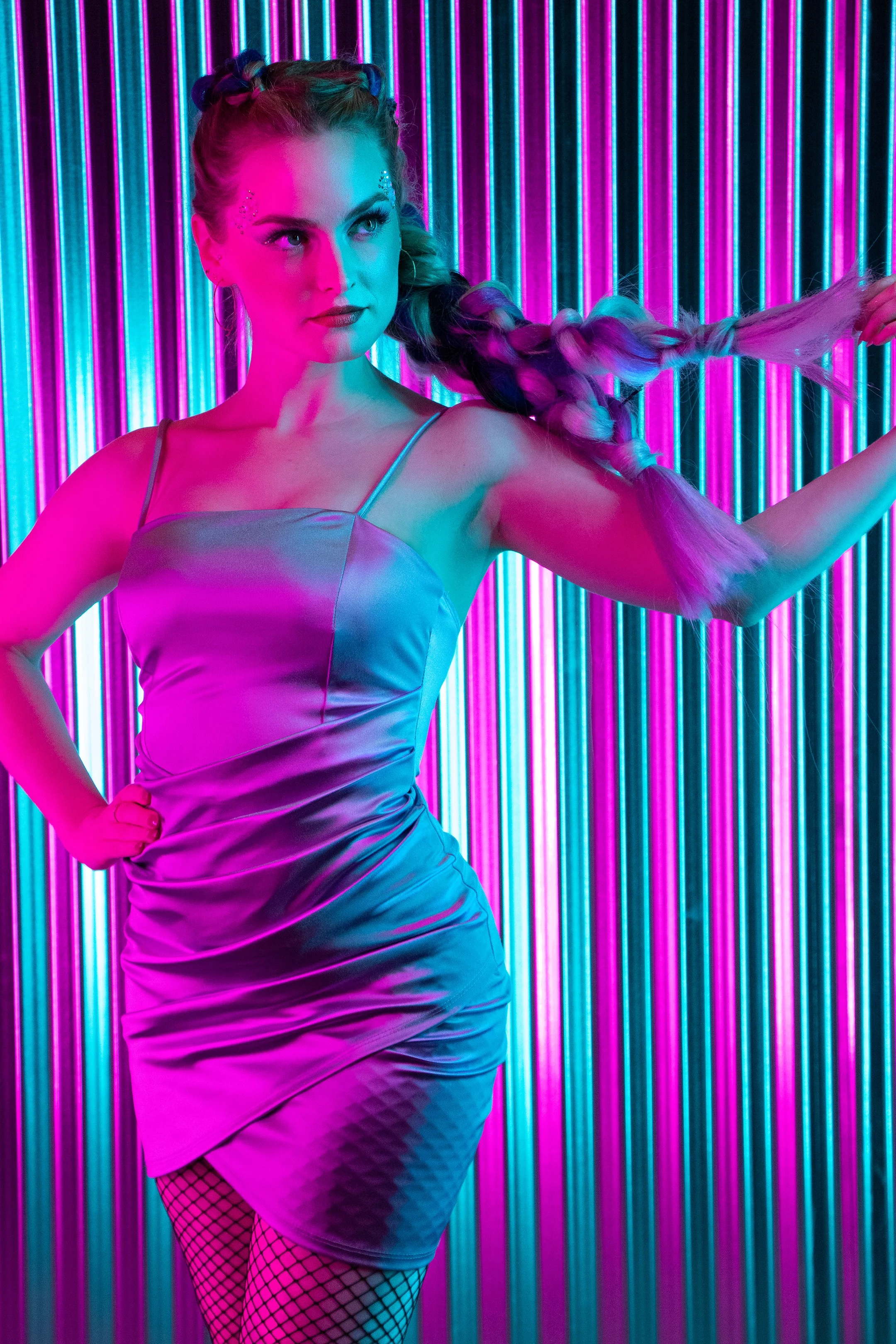 A woman with braided hair, glitter makeup, and fishnet stockings stands against a backdrop of colorful vertical neon lights, wearing a shiny satin dress.