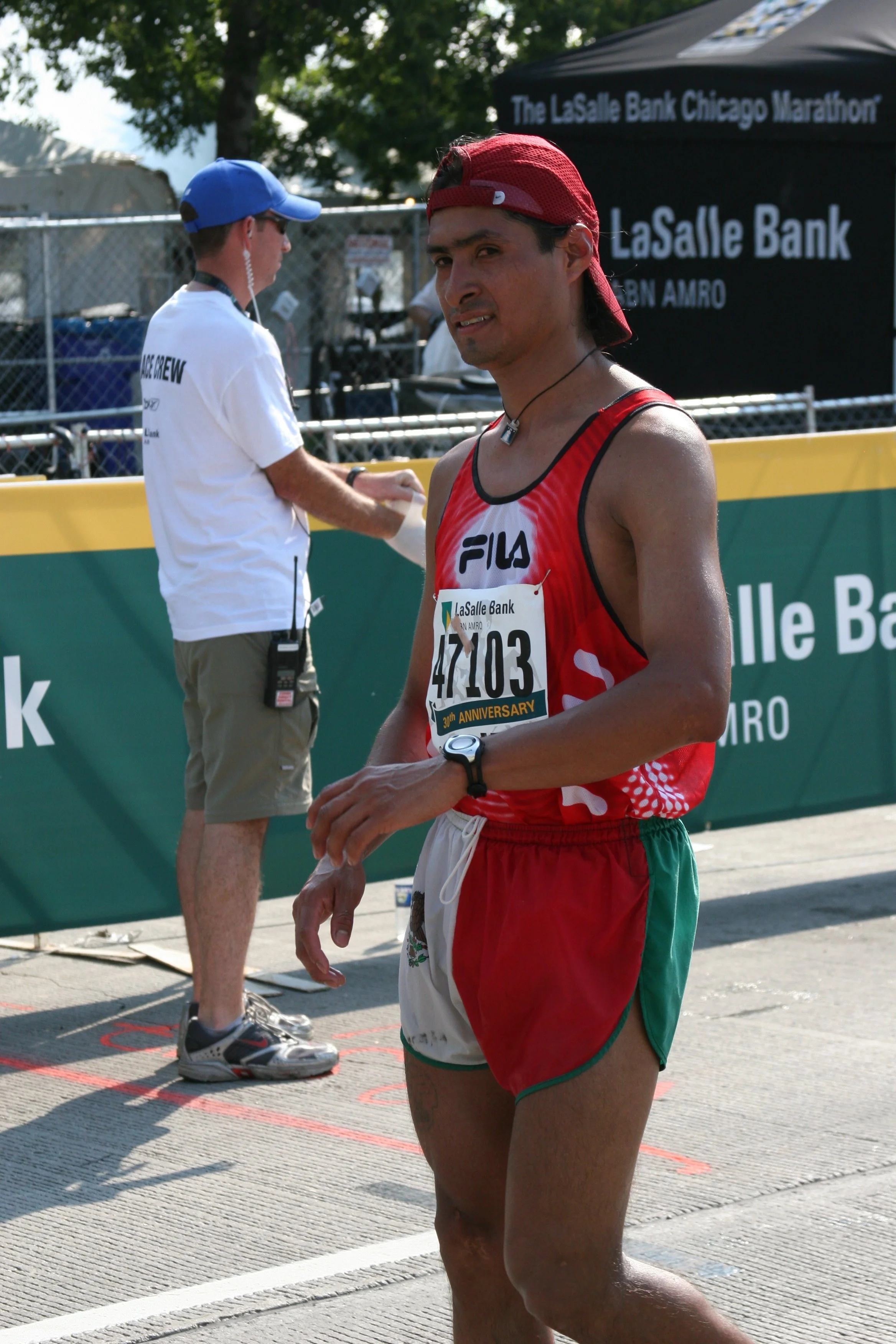 A male marathon runner in red and green running gear with a bib number, standing near the finish line at the Chicago Marathon, with a race official in a white shirt and blue cap in the background.