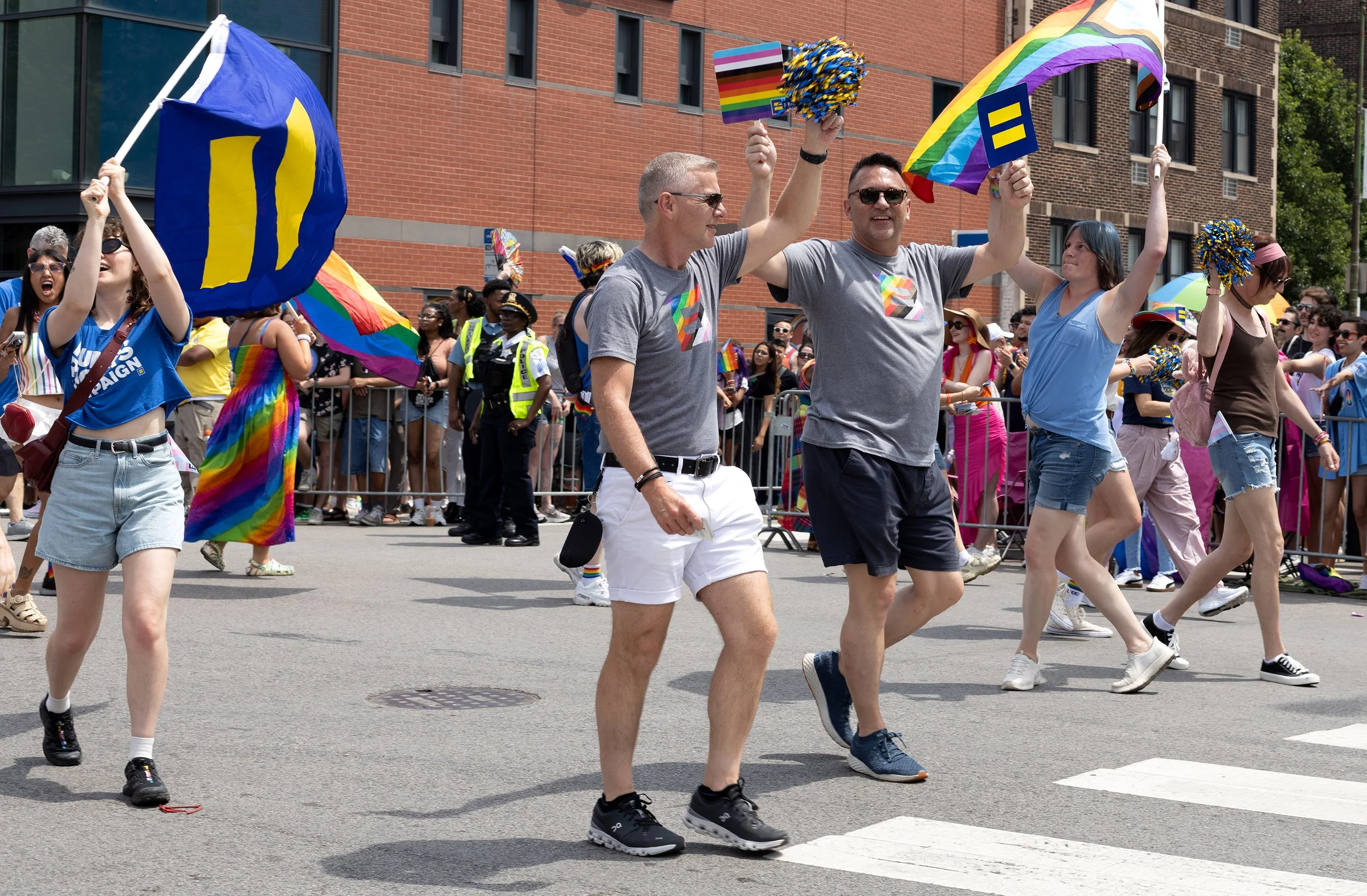People participating in a pride parade, holding rainbow flags and wearing colorful clothing and accessories, walking on a city street with a brick building in the background.