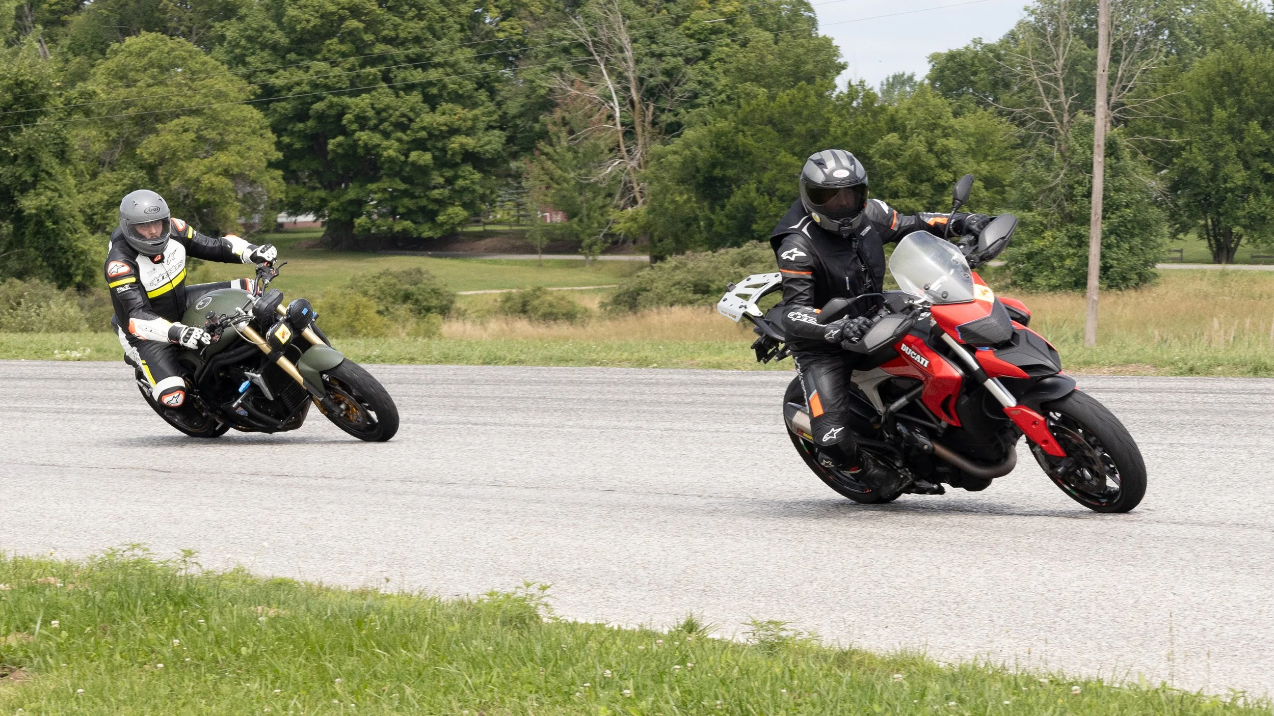 Two motorcyclists dressed in protective gear riding on a paved road surrounded by green grass and trees.
